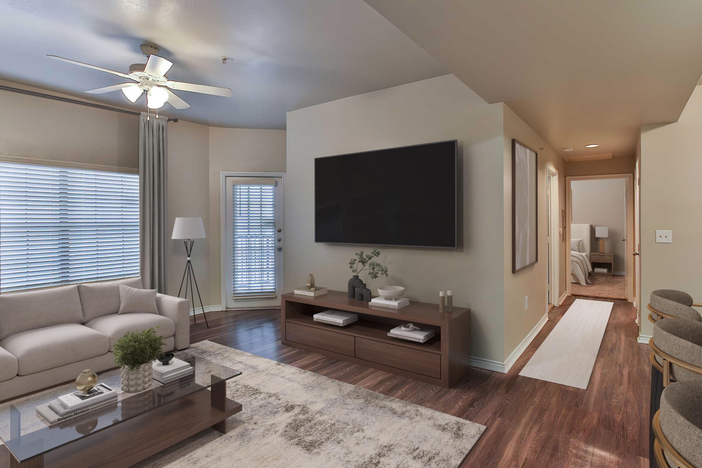 A modern living room featuring a beige sofa, a coffee table with decorative items, a large flat-screen TV mounted on the wall, and a fan on the ceiling. Natural light streams through the windows, and there is a door leading outside. Decor includes a rug, curtains, and a plant. An adjoining room is visible in the background.