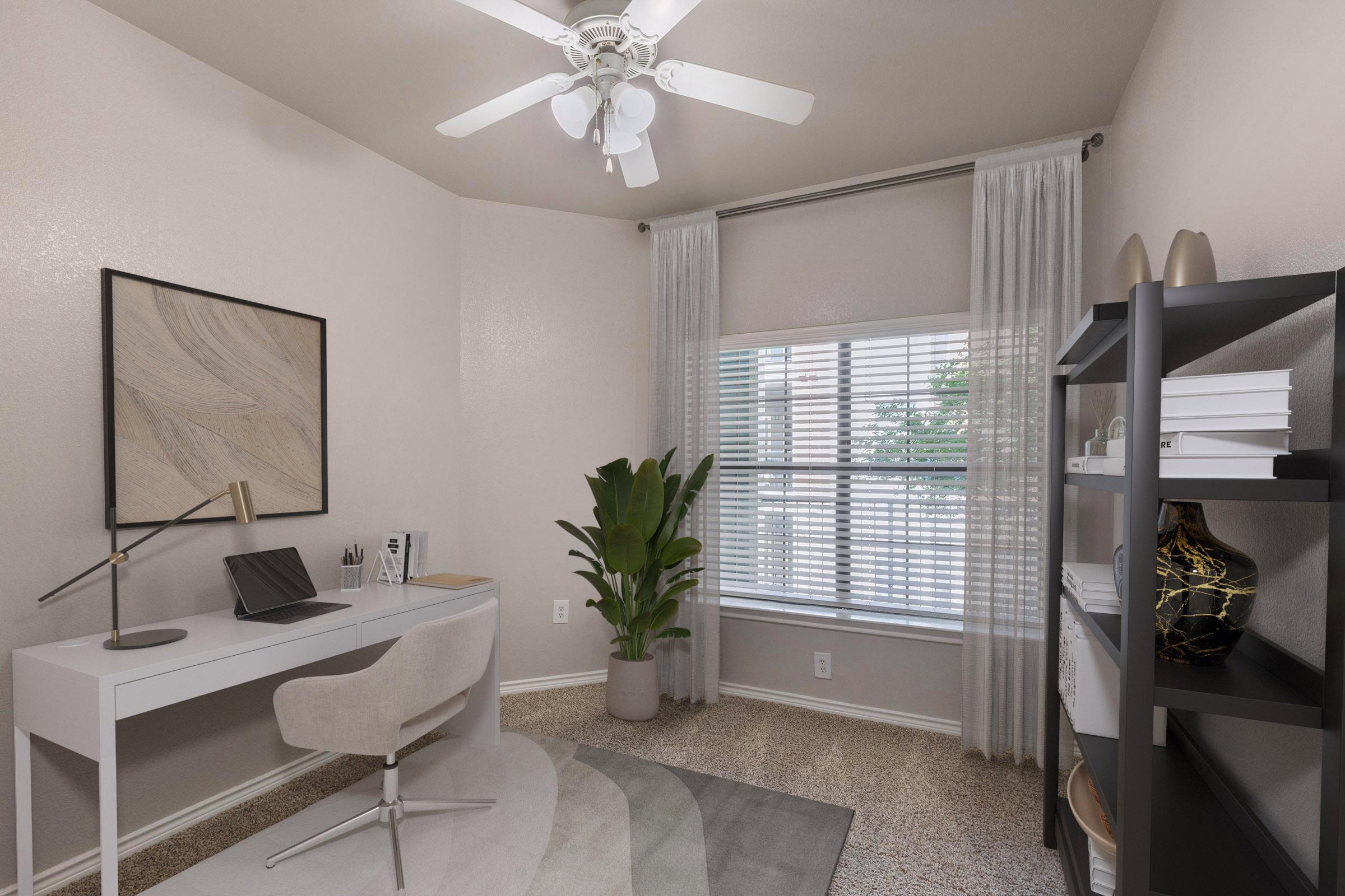 A modern home office featuring a white desk with a laptop, a comfortable chair, and decorative items. A tall potted plant adds a touch of greenery. The room has light-colored walls, a ceiling fan, and natural light streaming in through a window, which is framed by sheer curtains. A black bookshelf displays various items.