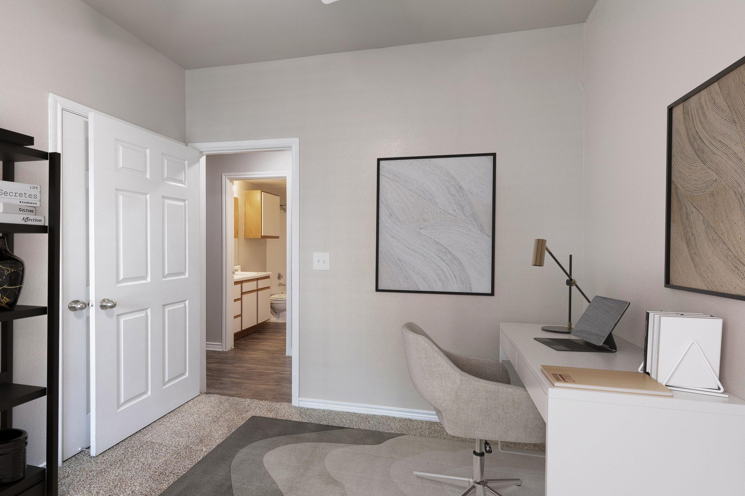 A cozy home office featuring a light-colored desk with a laptop and lamp, a comfortable chair, and a decorative wall art piece. There is a black shelving unit on the left and an open door leading to a bathroom in the background, with neutral-colored walls and flooring.