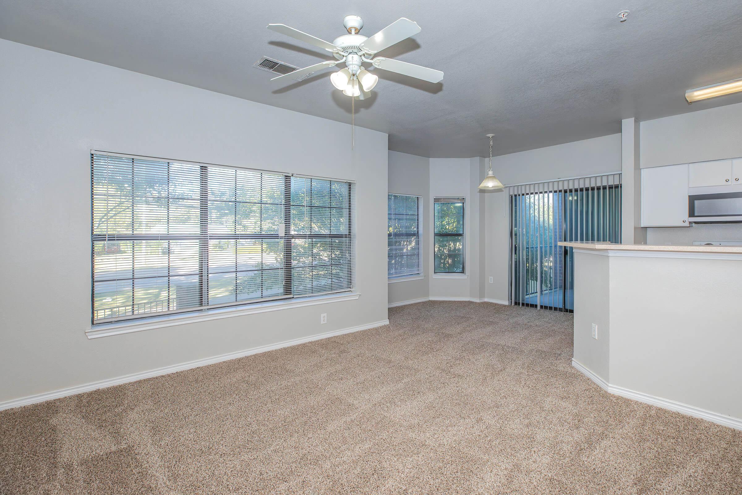 A spacious living room with light-colored walls and a ceiling fan. Large windows let in natural light, highlighting the plush carpet. A sliding glass door leads to a patio area. The adjacent kitchen is partially visible, with white cabinetry and modern appliances.