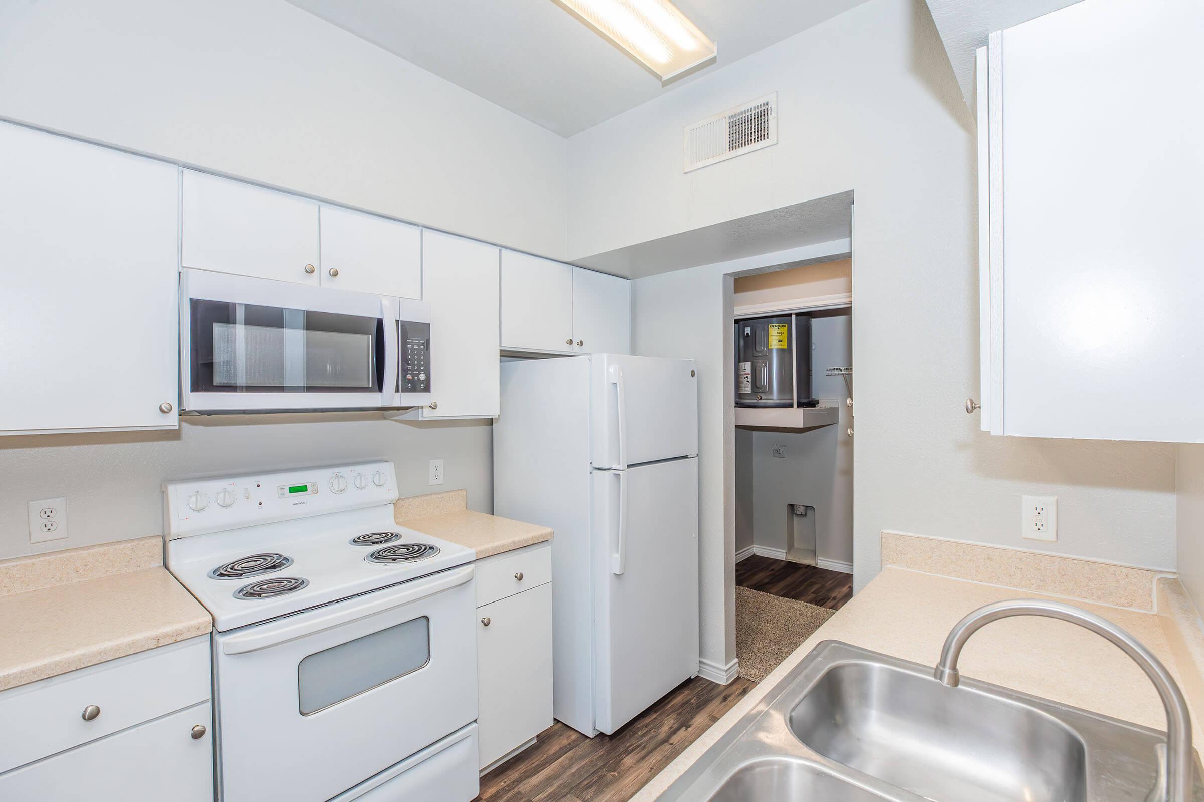 A modern kitchen featuring white cabinetry, a microwave, and a stove with an oven. There's a refrigerator and a double sink. The design includes light-colored countertops and wood-like flooring. A doorway leads to another area, indicating a well-organized and functional space.
