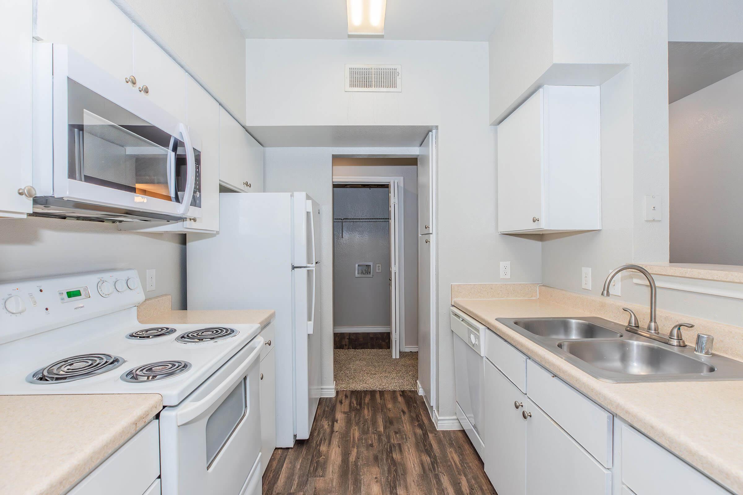 A bright and modern kitchen featuring white cabinetry, stainless steel sink, and appliances including a microwave and electric stove. The room has a light color palette with wood-like flooring and a doorway leading to a hallway with a closet.