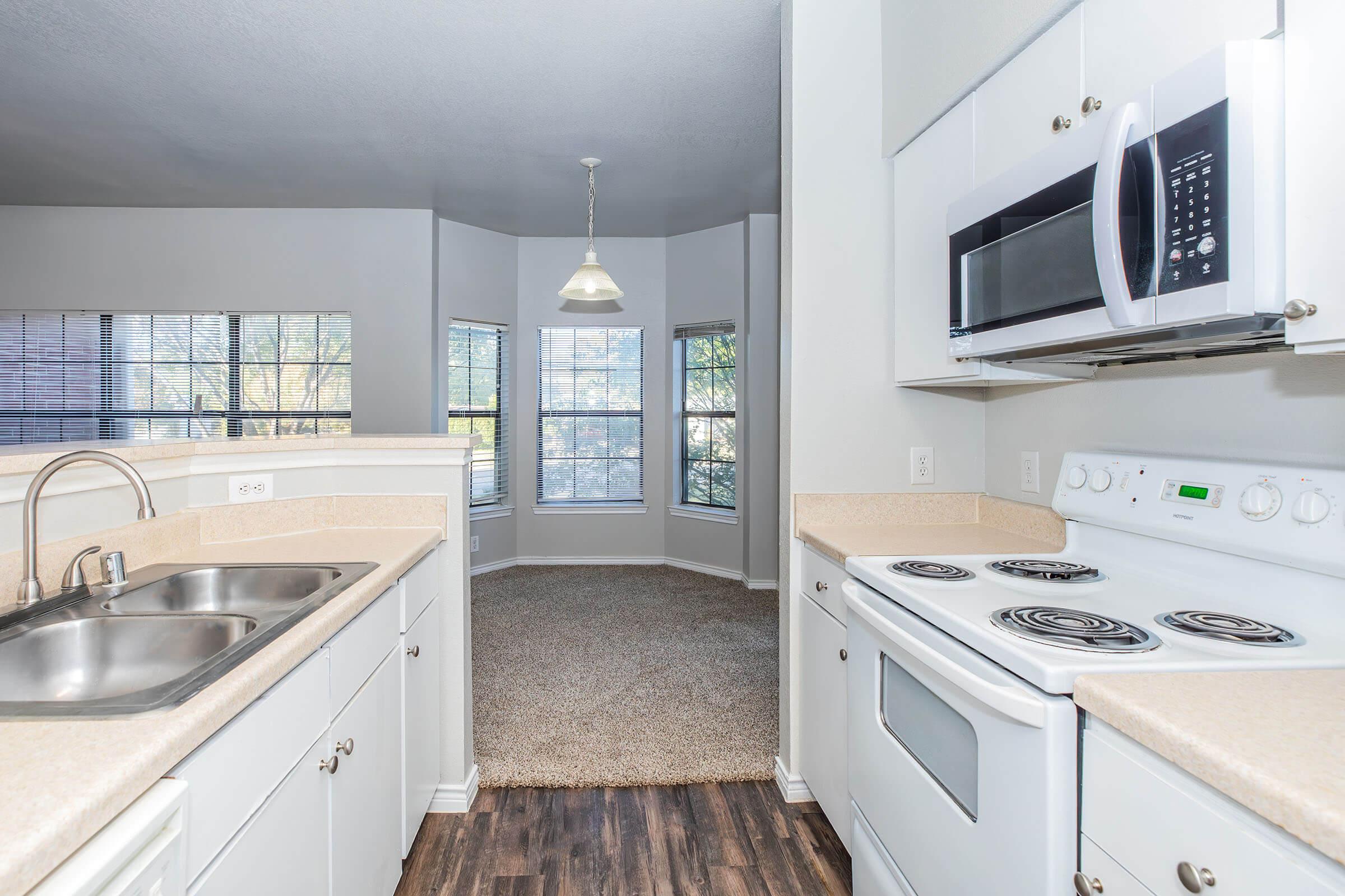 Bright and modern kitchen featuring a double sink, electric stove, and microwave, with cabinets and countertops in neutral tones. The frame opens to a cozy living area with large windows allowing natural light, showcasing carpeted flooring and a pendant light fixture overhead.
