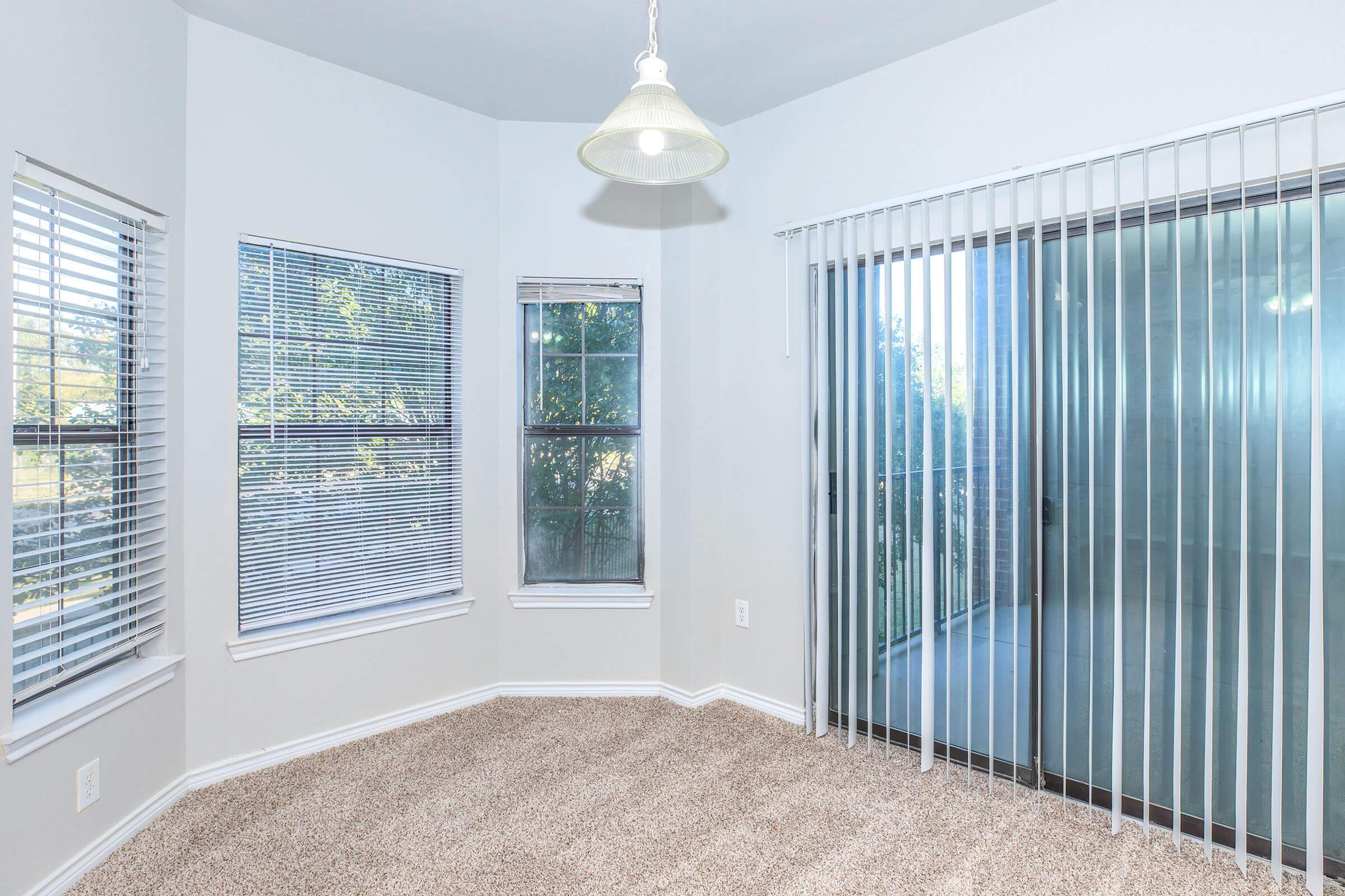 A well-lit corner of a room featuring beige carpet flooring. There are two windows with white blinds and a sliding glass door with vertical blinds that leads to a balcony. The walls are painted light gray, and a modern pendant light hangs from the ceiling, enhancing the bright and airy feel of the space.