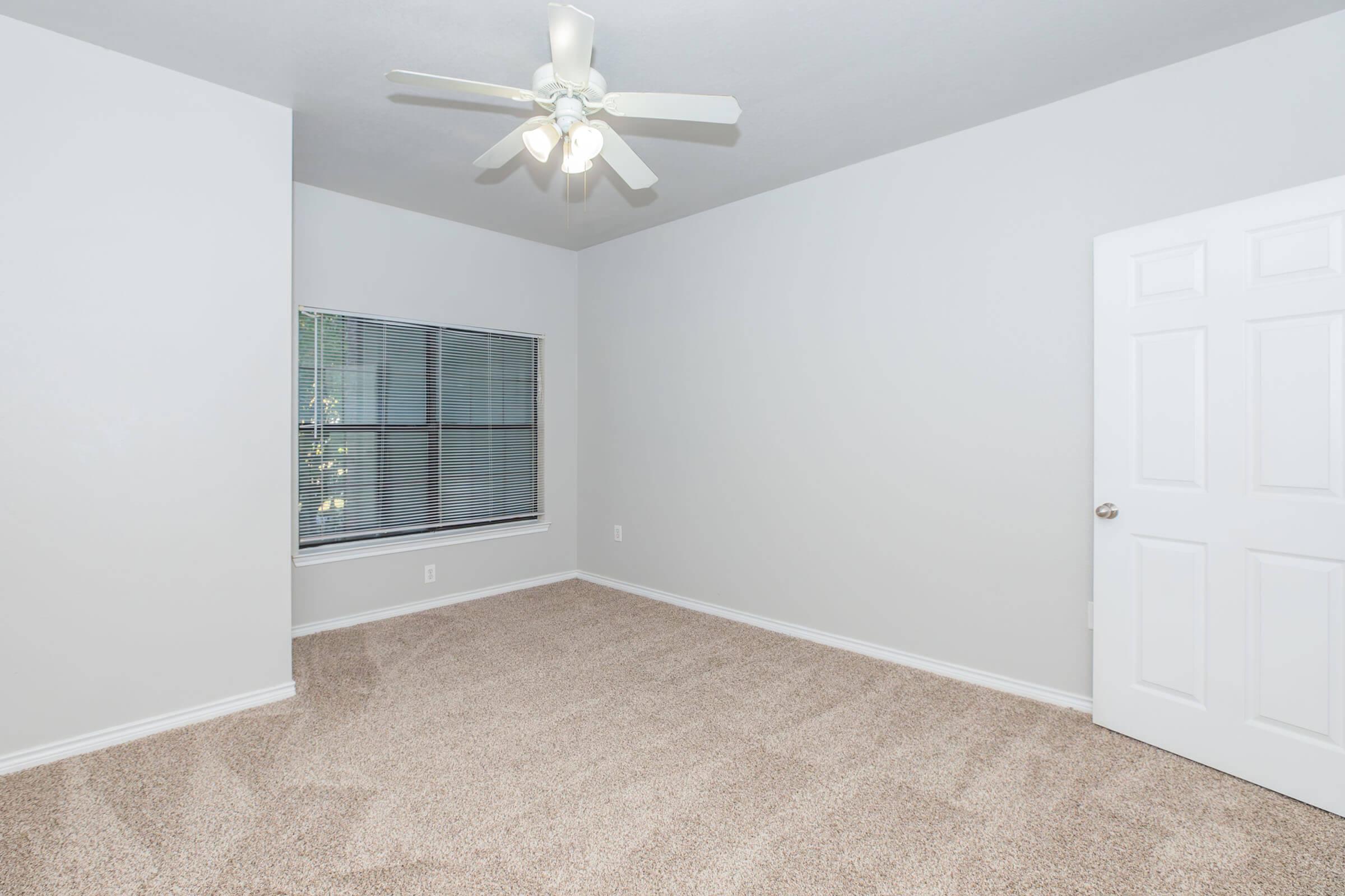 A spacious, empty room with light gray walls and a ceiling fan. There is a large window with blinds letting in natural light, and the floor is covered in plush, light-colored carpeting. A white door is visible on the right side of the image.