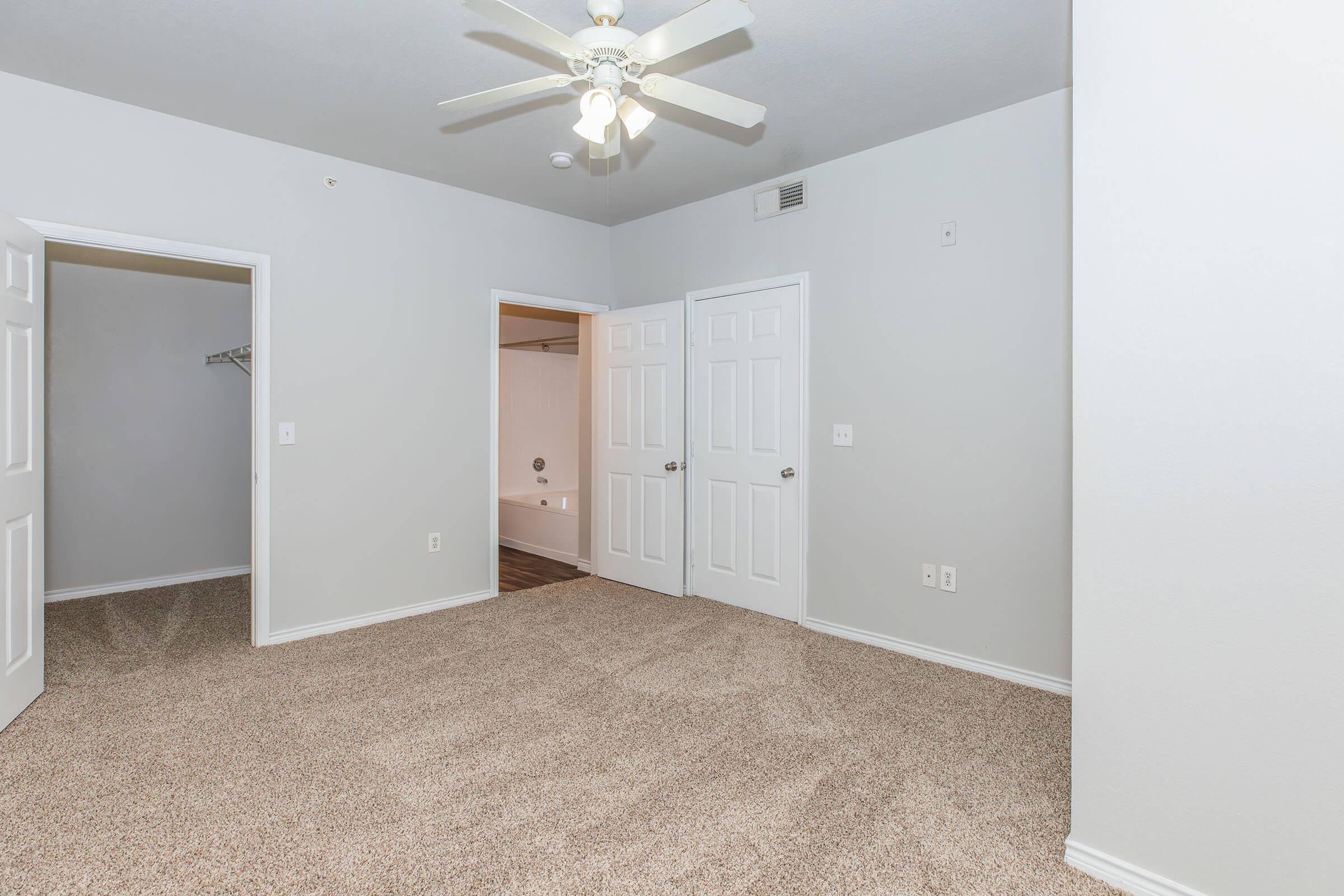 A spacious room with a light gray wall color and carpeted flooring. There is a ceiling fan overhead. Two closed doors lead to adjacent areas, one of which has a small closet. The room appears bright and well-lit, with a neutral aesthetic conducive to personalization.