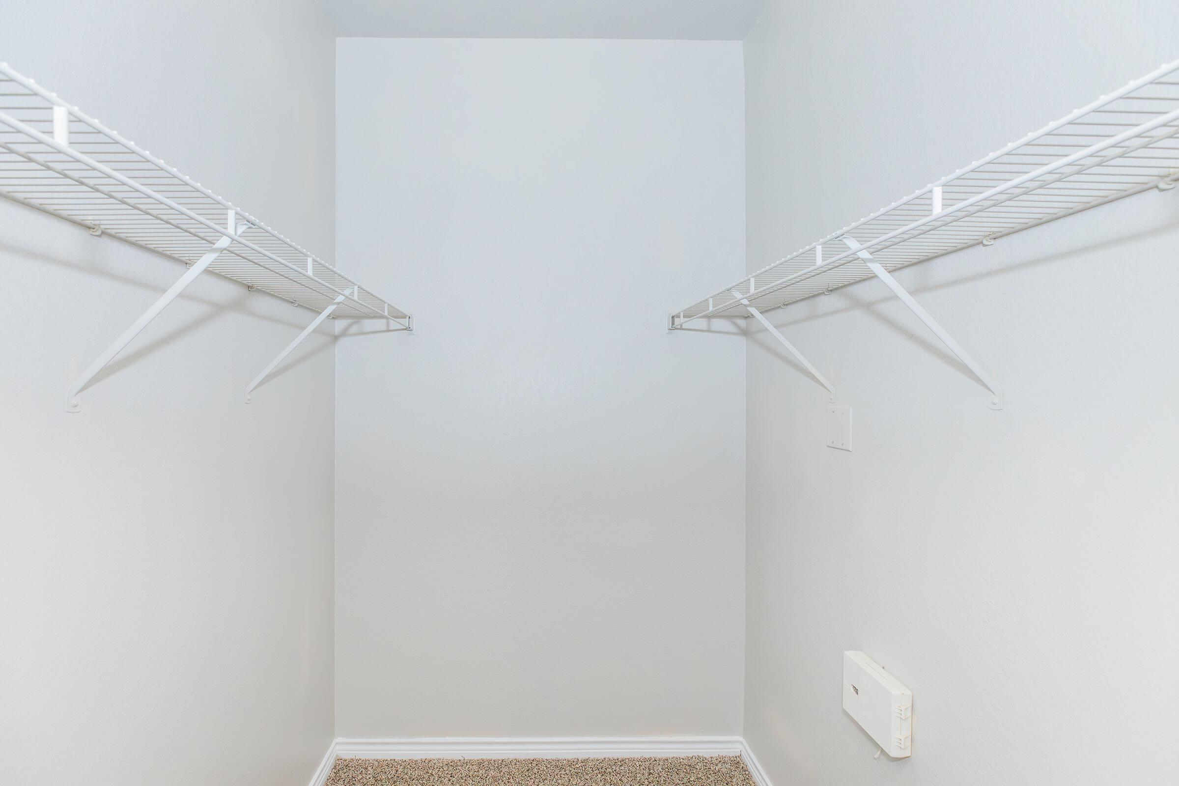 Empty closet with white walls and two metal wire shelves mounted on either side. The floor is carpeted, and there is a small white wall-mounted fixture on the right side. The space is well-lit and organized, highlighting its minimalistic design.