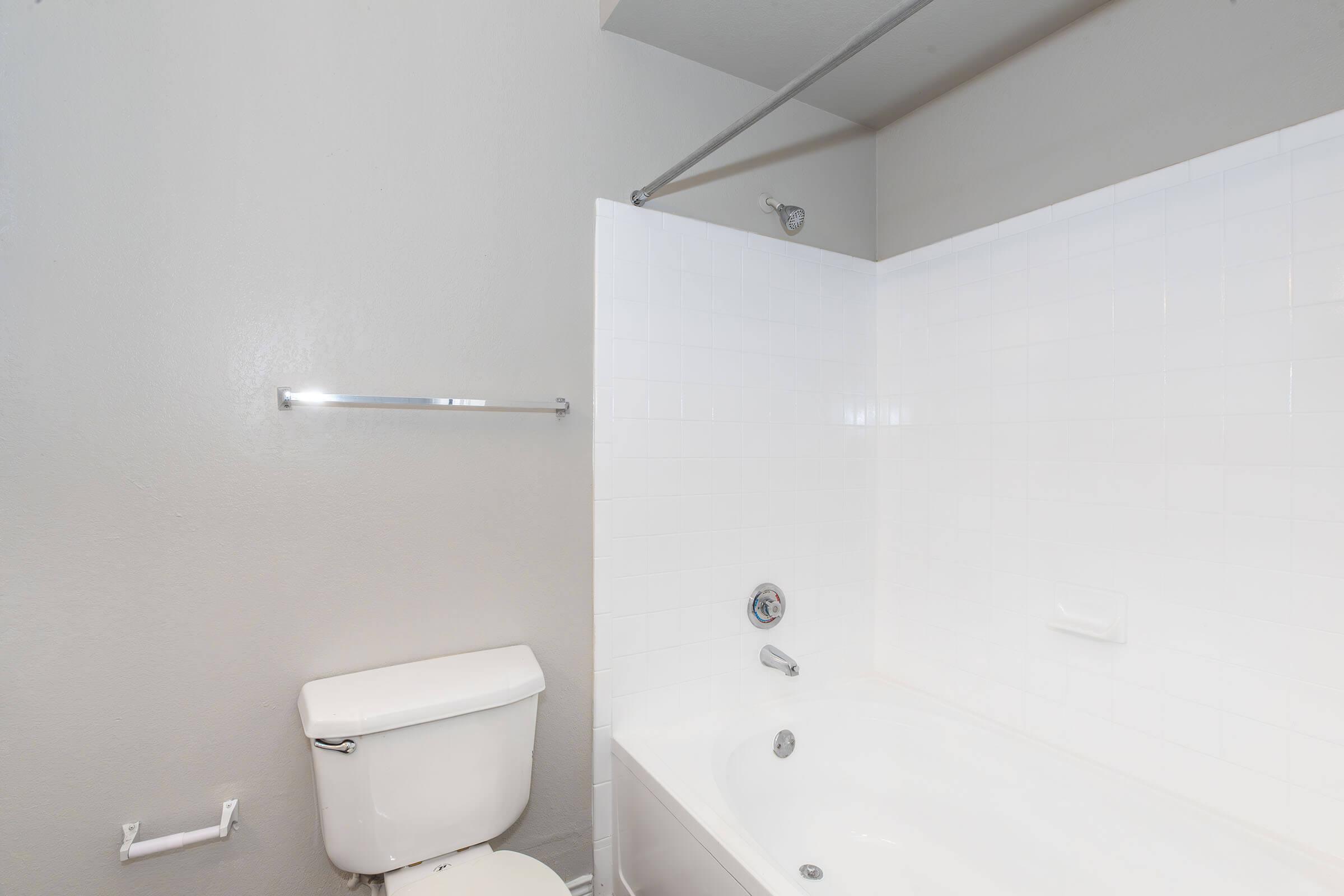A bathroom with a white bathtub and shower combination, a white toilet, and gray walls. There is a towel bar mounted on the wall and a faucet visible in the bathtub area. The space appears clean and well-lit, featuring simple, modern design elements.