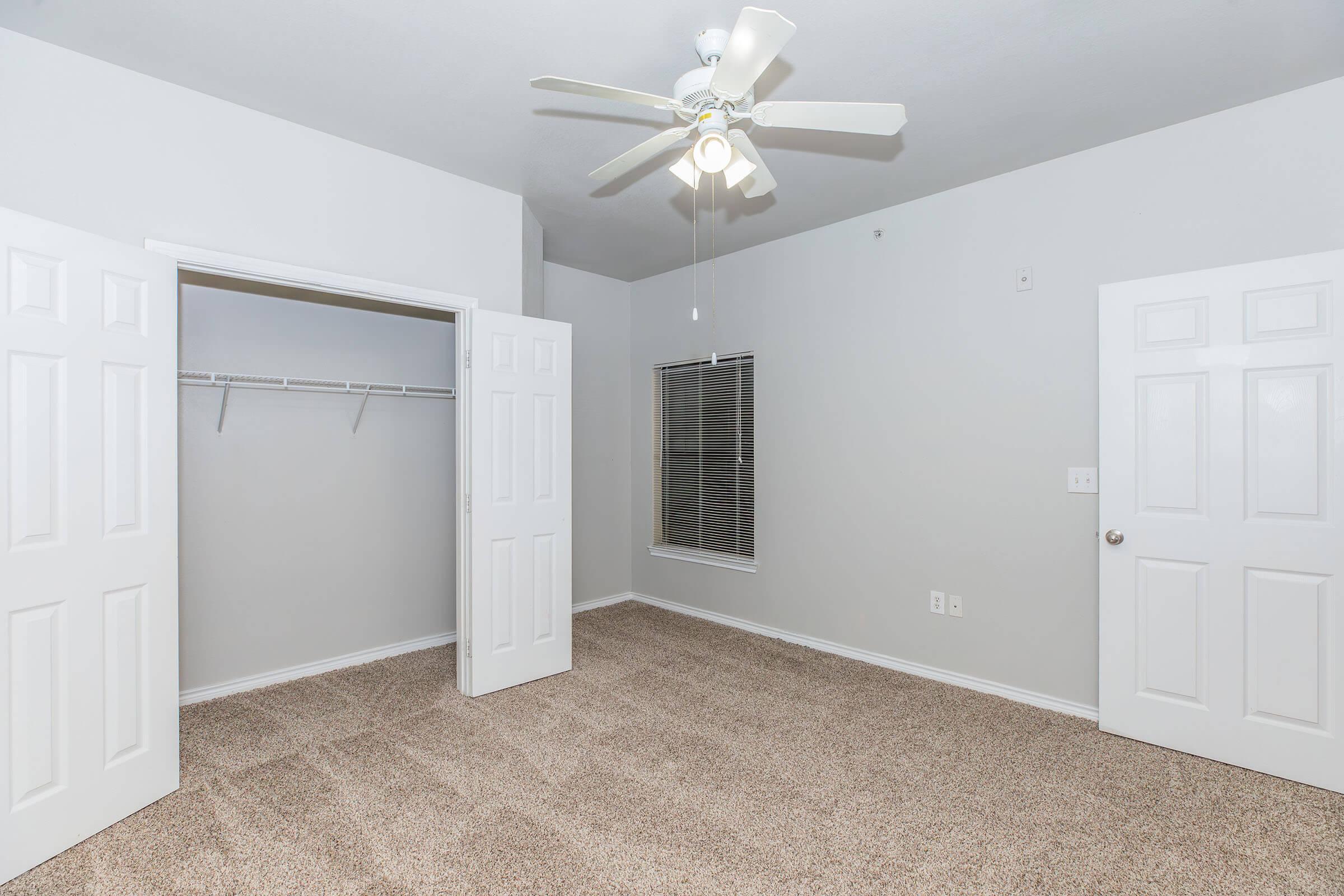 A brightly lit, empty bedroom featuring light gray walls and plush beige carpeting. The room includes a ceiling fan, a window with blinds, and two doors—one leading to a closet and another possibly to a hallway.