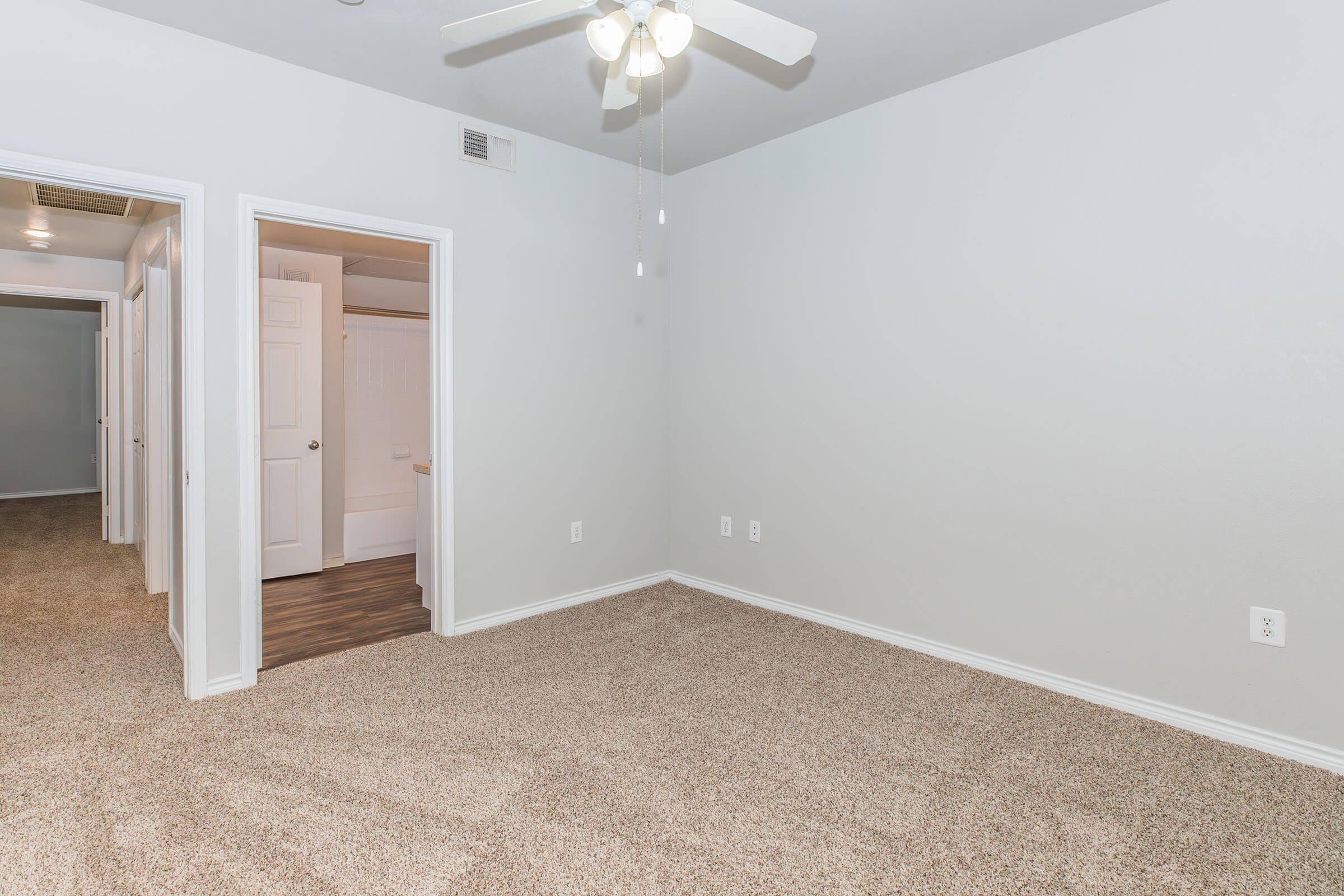 A spacious, empty room featuring light gray walls and beige carpet flooring. A ceiling fan is mounted in the center, and there are two doors visible: one leading to a bathroom and the other to a hallway. The room has a bright and airy feel, with ample natural light coming through the open doors.