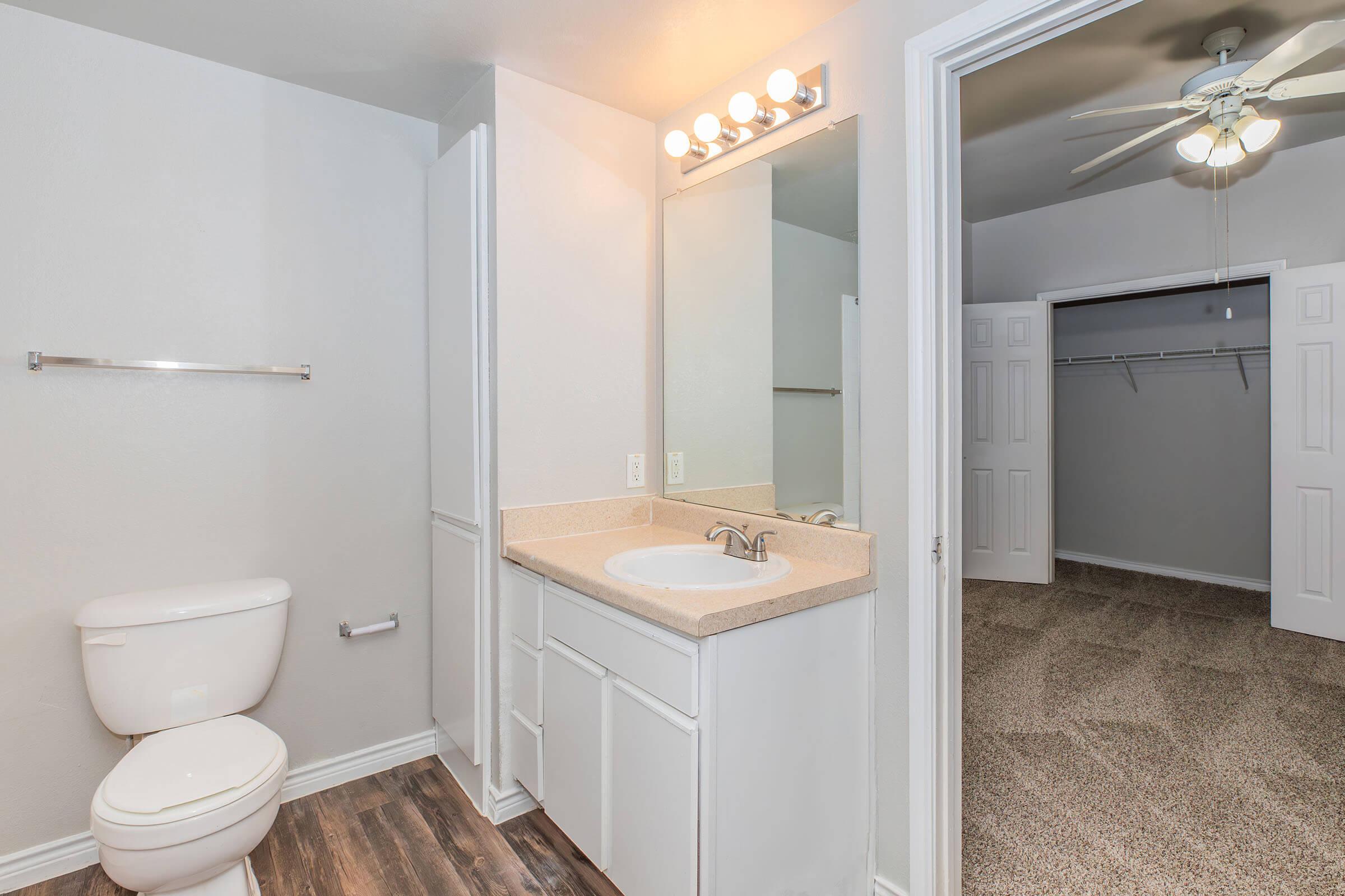 A clean, modern bathroom featuring a white toilet, a light-colored vanity with a sink and mirror above, and an adjacent door leading to a carpeted room with a ceiling fan. The walls are painted light gray, and there is a towel bar on the wall next to the vanity.