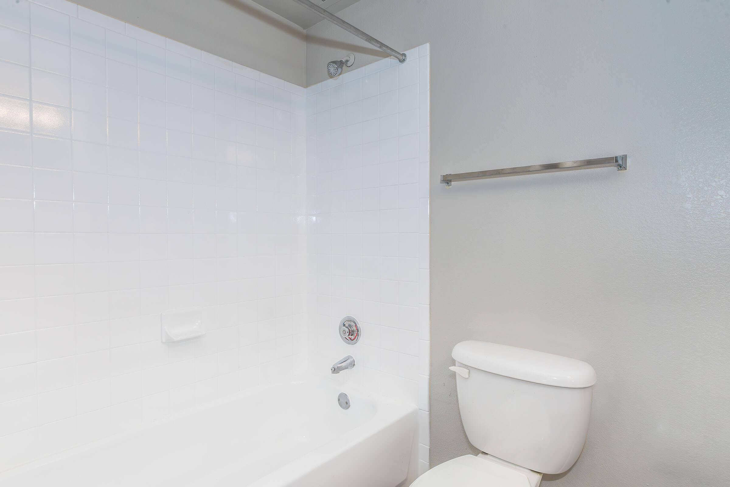 A clean and modern bathroom featuring a white tiled bathtub with a silver faucet, a toilet, and a towel bar. The walls are painted a light gray, providing a neutral backdrop. Natural light enhances the bright, minimalist aesthetic.