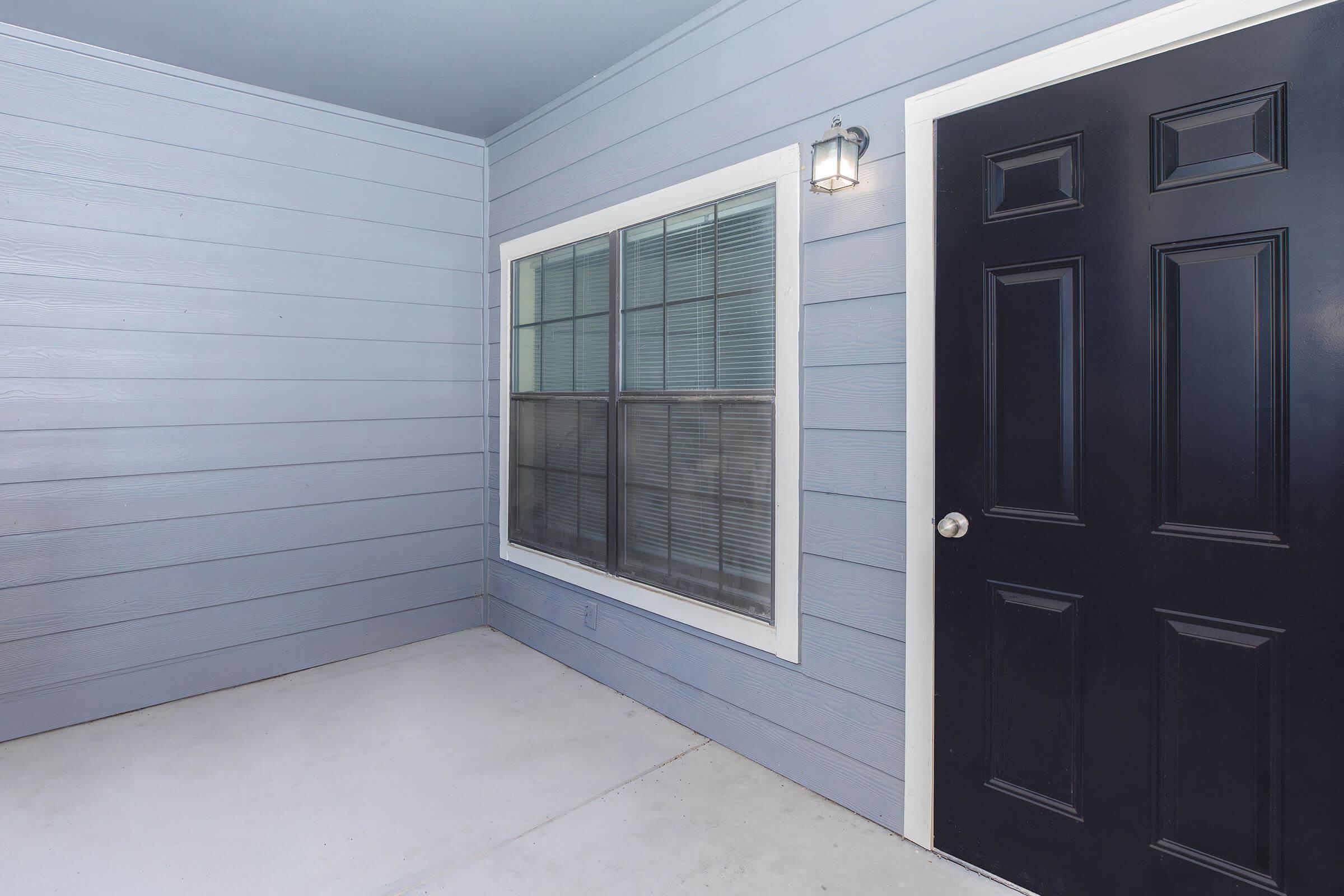 Exterior view of a modern entryway featuring a black door with a round knob, a large window beside it with multiple panes, and light blue wooden siding. The scene is well-lit, showcasing a clean and inviting space.