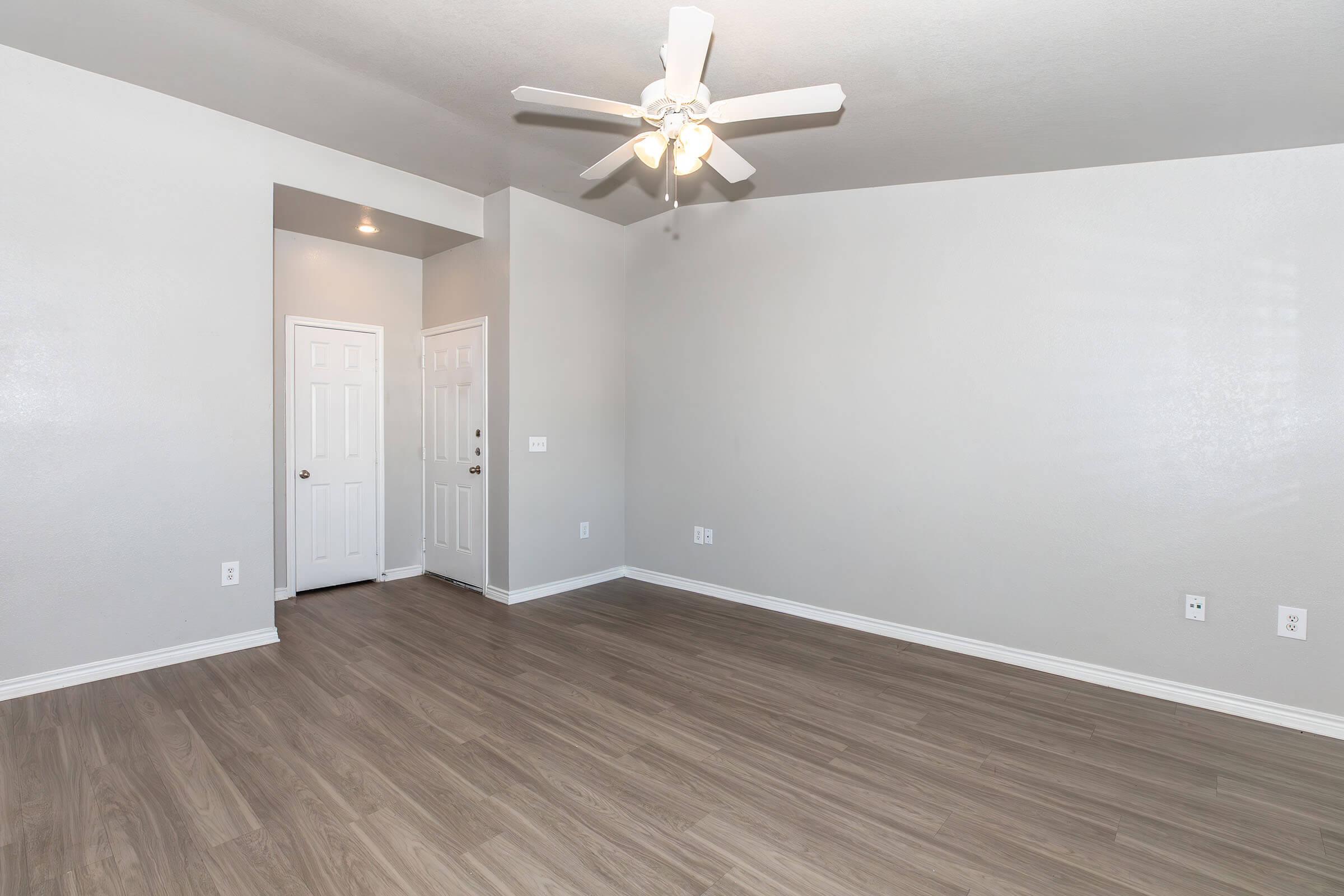 A spacious, empty room with light gray walls and a light wood laminate floor. A ceiling fan is mounted in the center, and there are two white doors visible on the left side. The overall atmosphere is bright and inviting, with ample space for furniture placement.