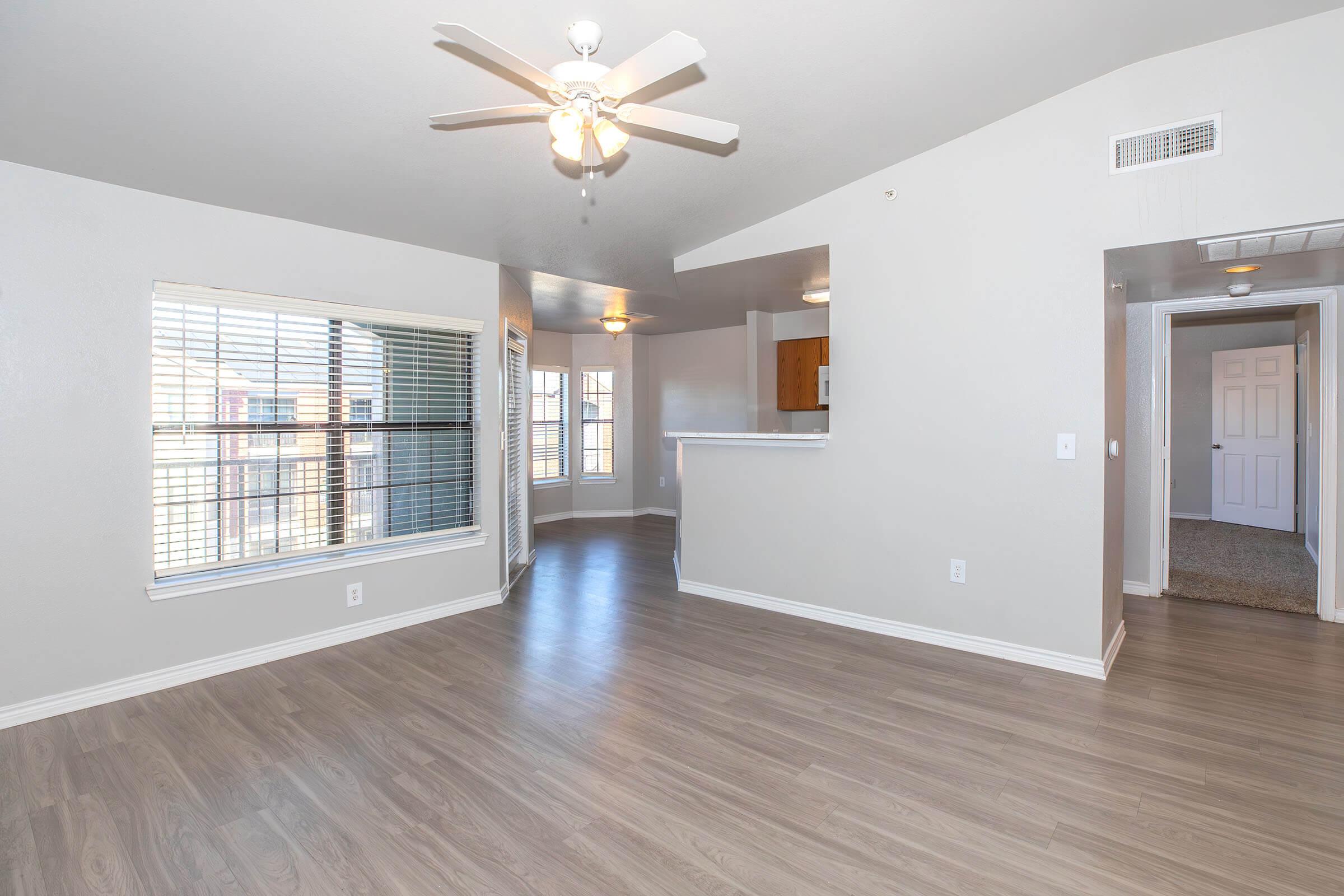 Spacious living room featuring a ceiling fan, large windows that allow natural light, and light-colored walls. The floor is finished with sleek hardwood-like flooring, and there's an open view toward a kitchen area. Doorways lead to adjacent rooms, enhancing the open layout.