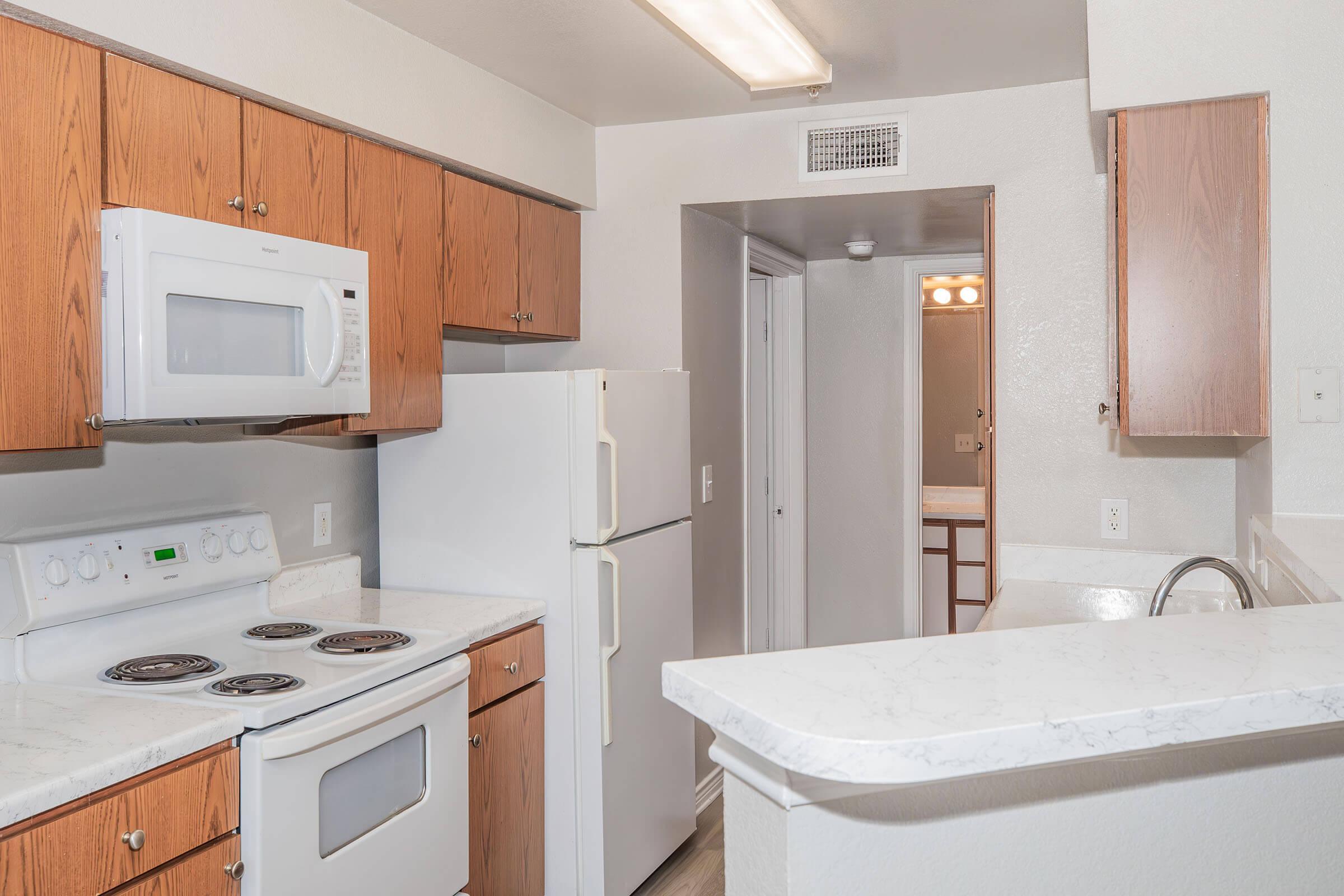 A modern kitchen featuring wooden cabinets, white appliances including a microwave and refrigerator, a stove with four burners, and a countertop. The space has bright lighting and an open layout leading to a hallway.