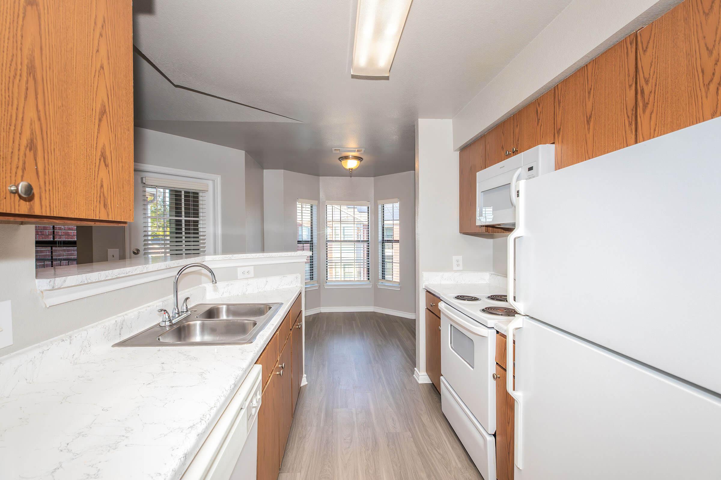 Bright and modern kitchen featuring wooden cabinetry, a double sink, white appliances including an oven and refrigerator, and a marble countertop. Natural light fills the space from multiple windows, and the flooring is sleek and wood-like.