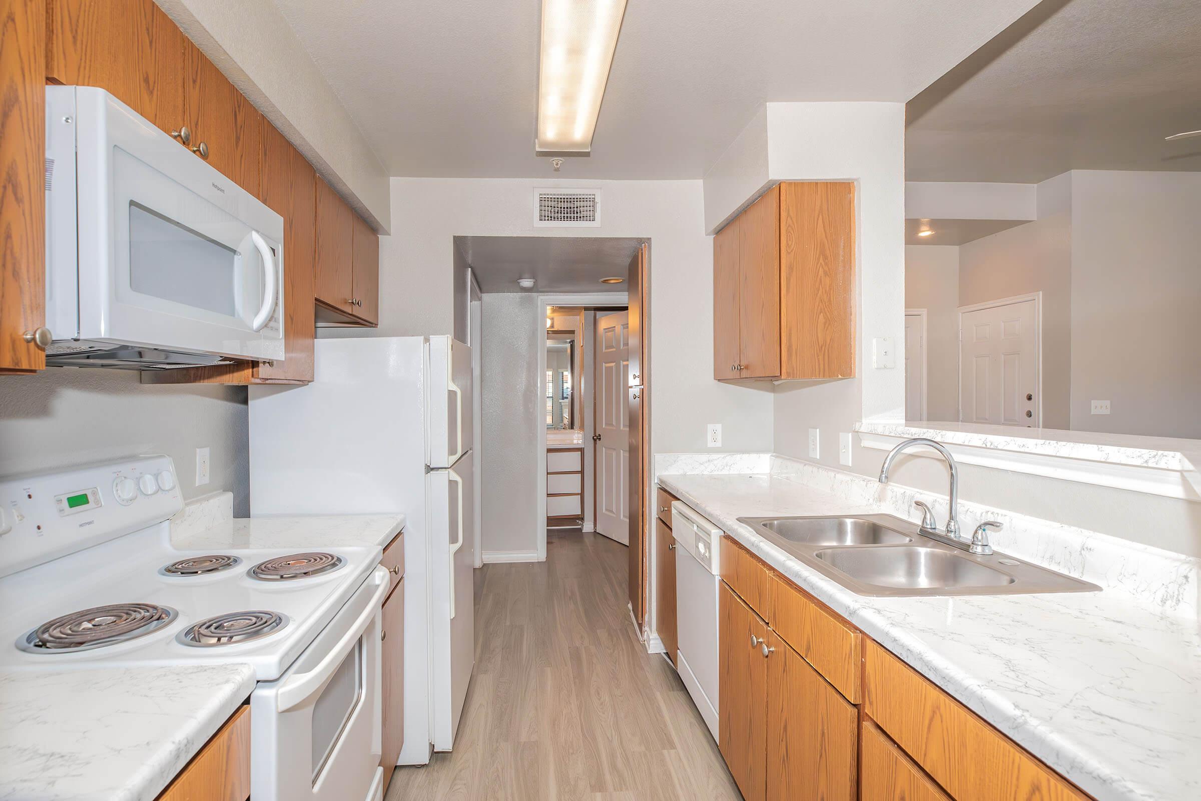 Modern kitchen with wooden cabinets, white appliances including a microwave and refrigerator, beige countertops, and a double sink. The layout features a narrow hallway leading to another room, enhancing the open feel of the space. Neutral colors create a warm and inviting atmosphere.