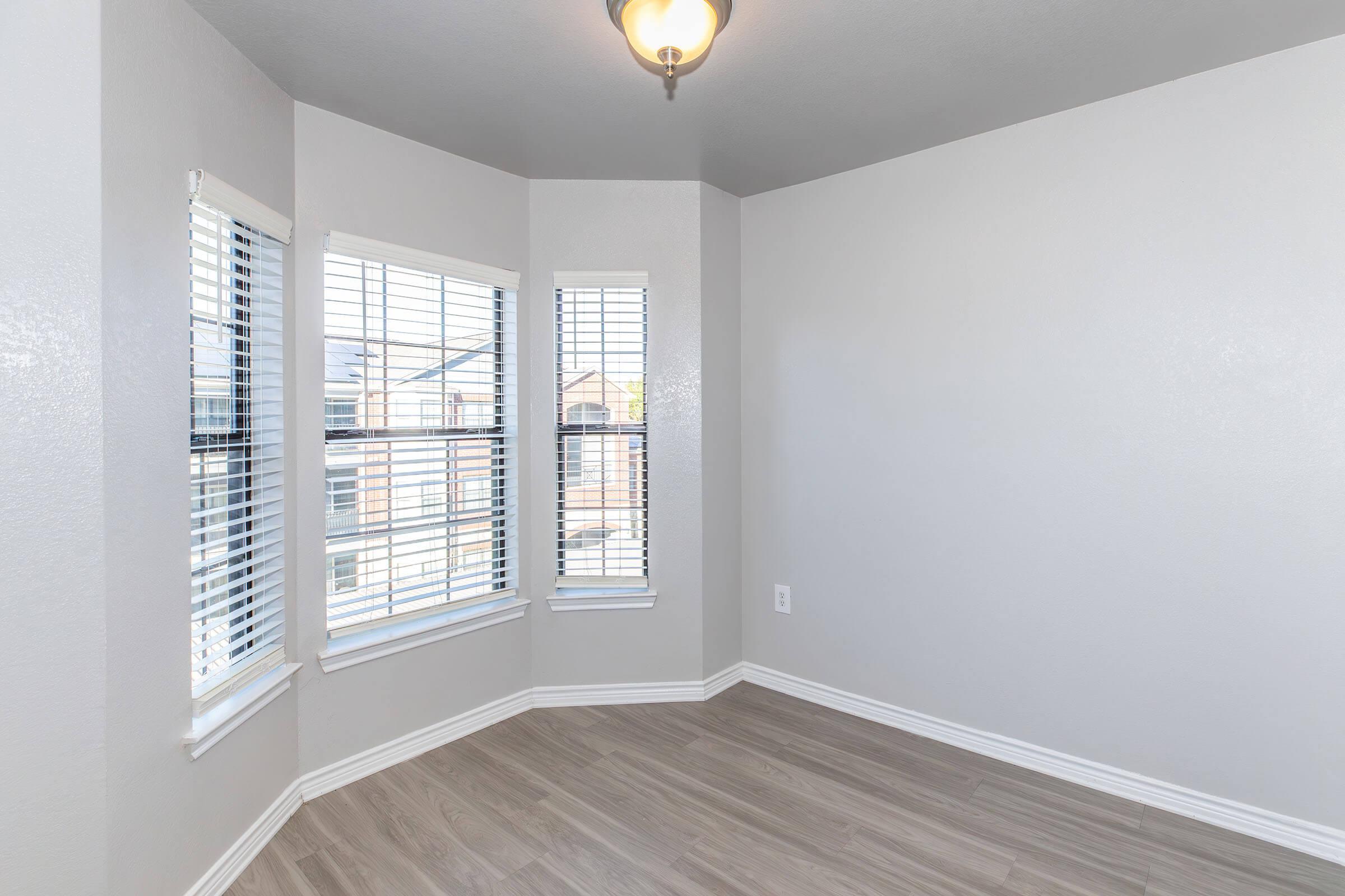 A bright, empty room with light gray walls and a light wood floor. There are three windows with white blinds allowing natural light to enter. The ceiling features a light fixture, and the room has a modern, clean appearance, suitable for various uses.