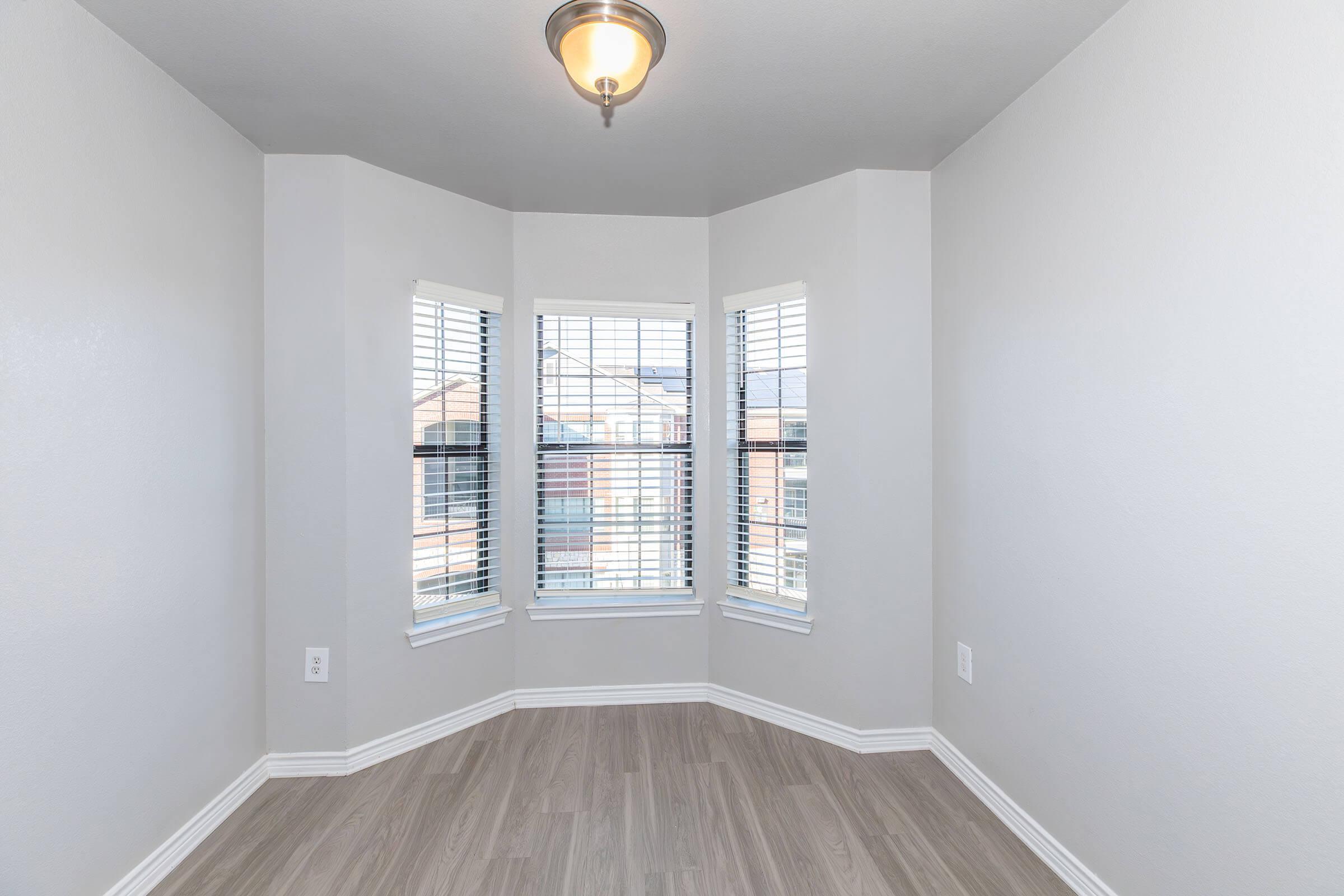 A bright, empty room with light gray walls and a wooden floor. The space features three large windows with white blinds, allowing natural light to enter. A modern ceiling light fixture is present, highlighting the clean and simple design.