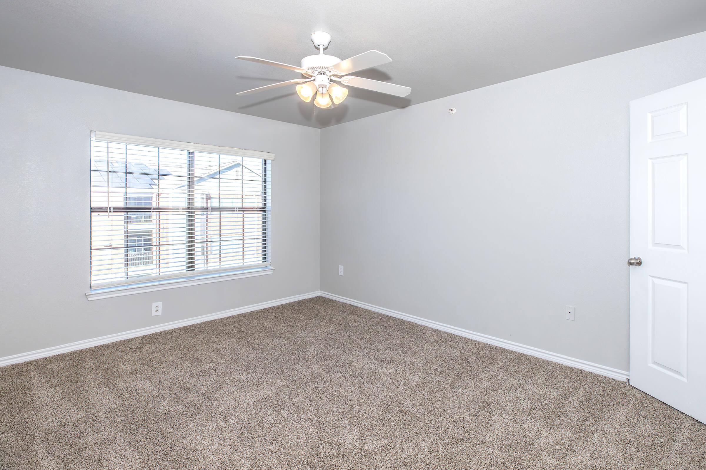 A spacious, empty room with light gray walls and plush carpet. It features a ceiling fan with four lights, a window with white blinds allowing natural light, and a white door, creating a bright and inviting atmosphere.