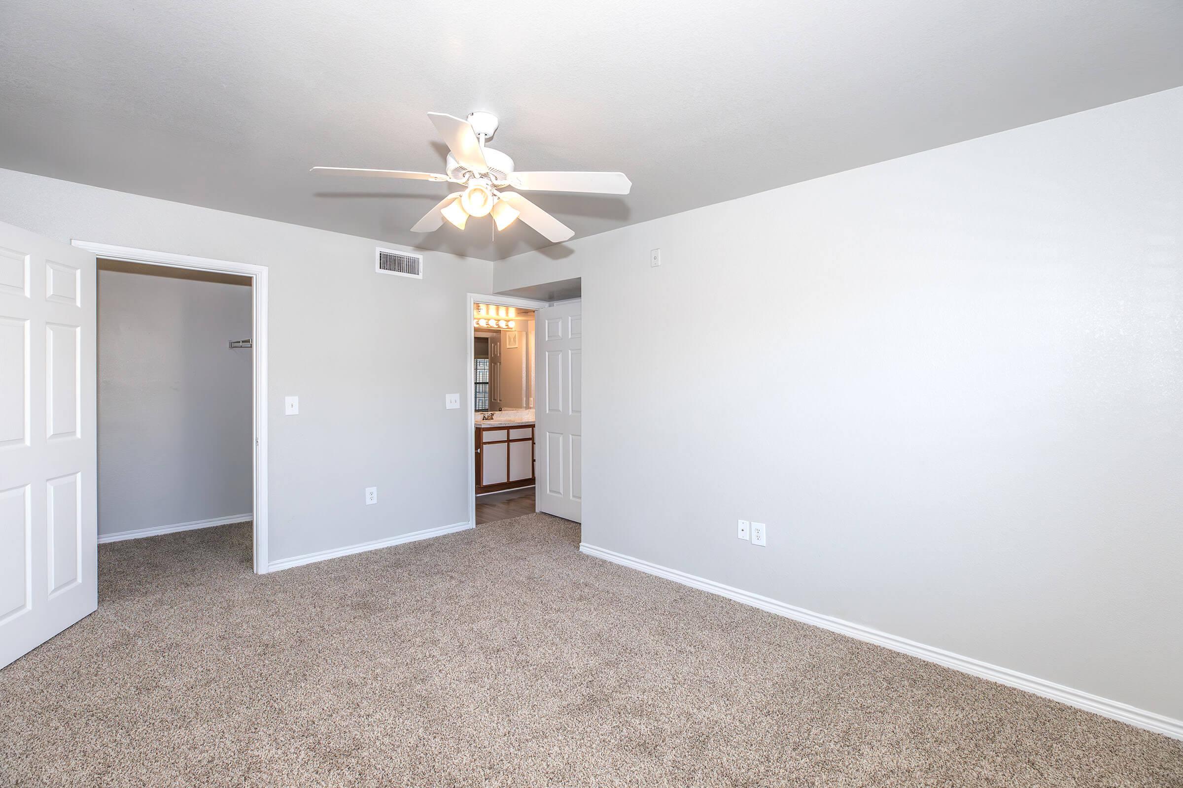 A spacious room with light beige carpeting and light gray walls, featuring a ceiling fan. There are two closed doors, one leading to a closet and another to a bathroom area in the background. Natural light brightens the space, giving it a cozy and inviting feel.