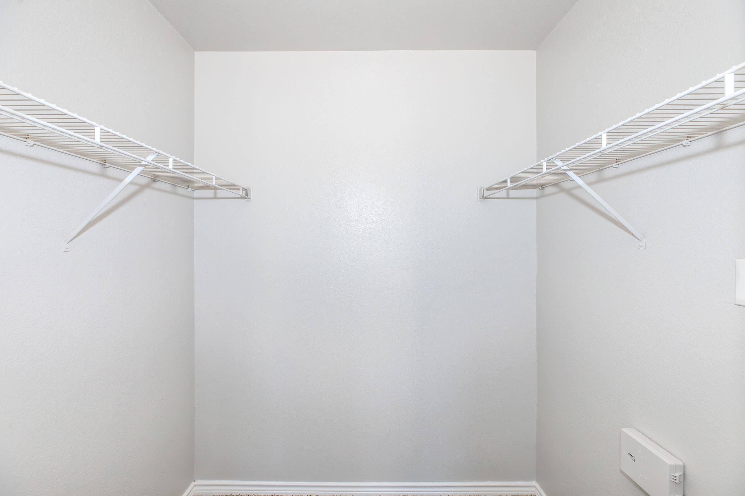 Empty closet with white walls and two wire shelving units on opposite sides, creating an open space. The floor is bare, and there is a small electrical outlet visible on the right wall. The overall appearance is clean and uncluttered.