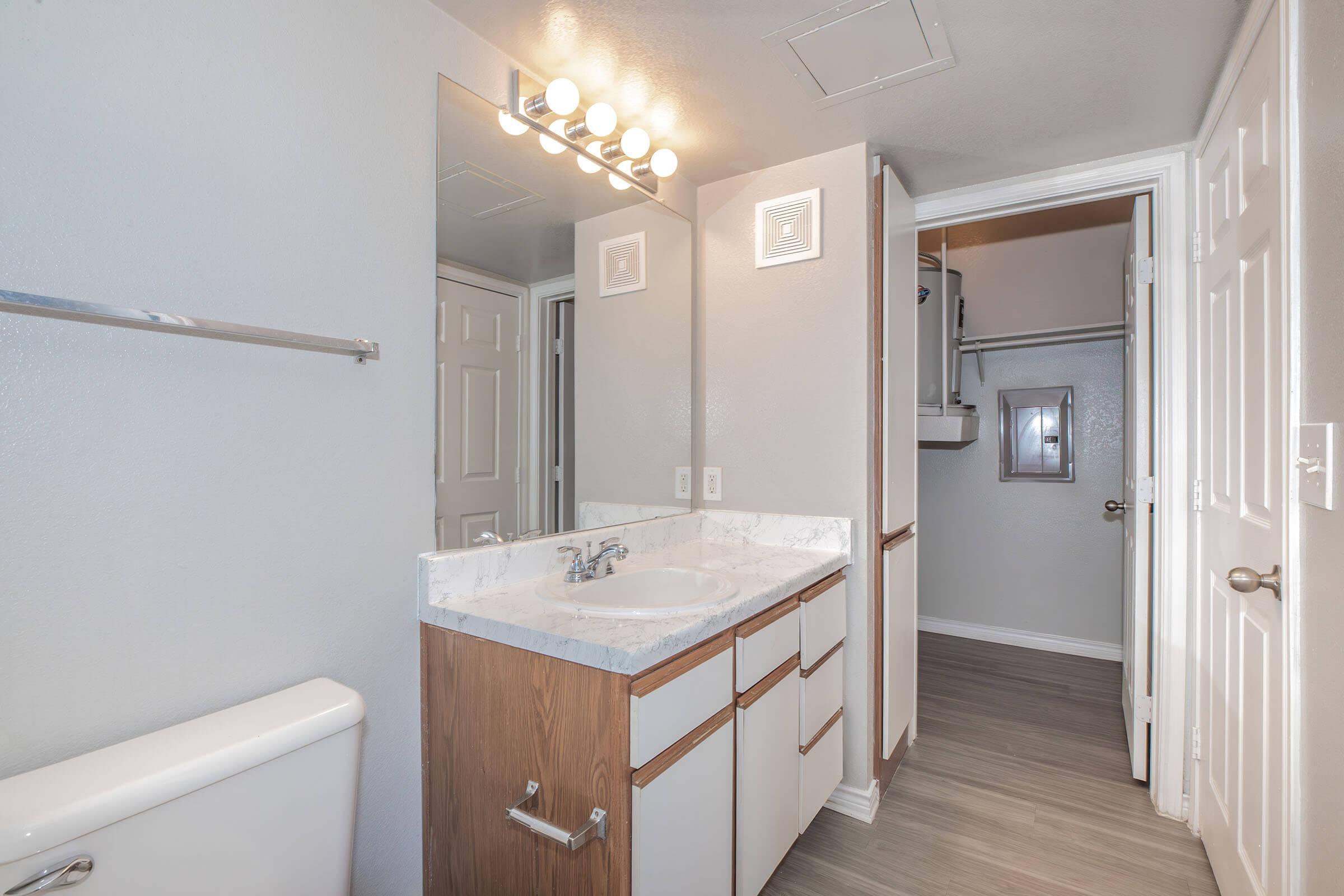 Modern bathroom interior featuring a double sink vanity with a white marble countertop and wooden cabinetry. A large mirror is above the sink. The bathroom includes a toilet and accessible closet space, with neutral gray walls and laminate flooring. Good lighting enhances the space.