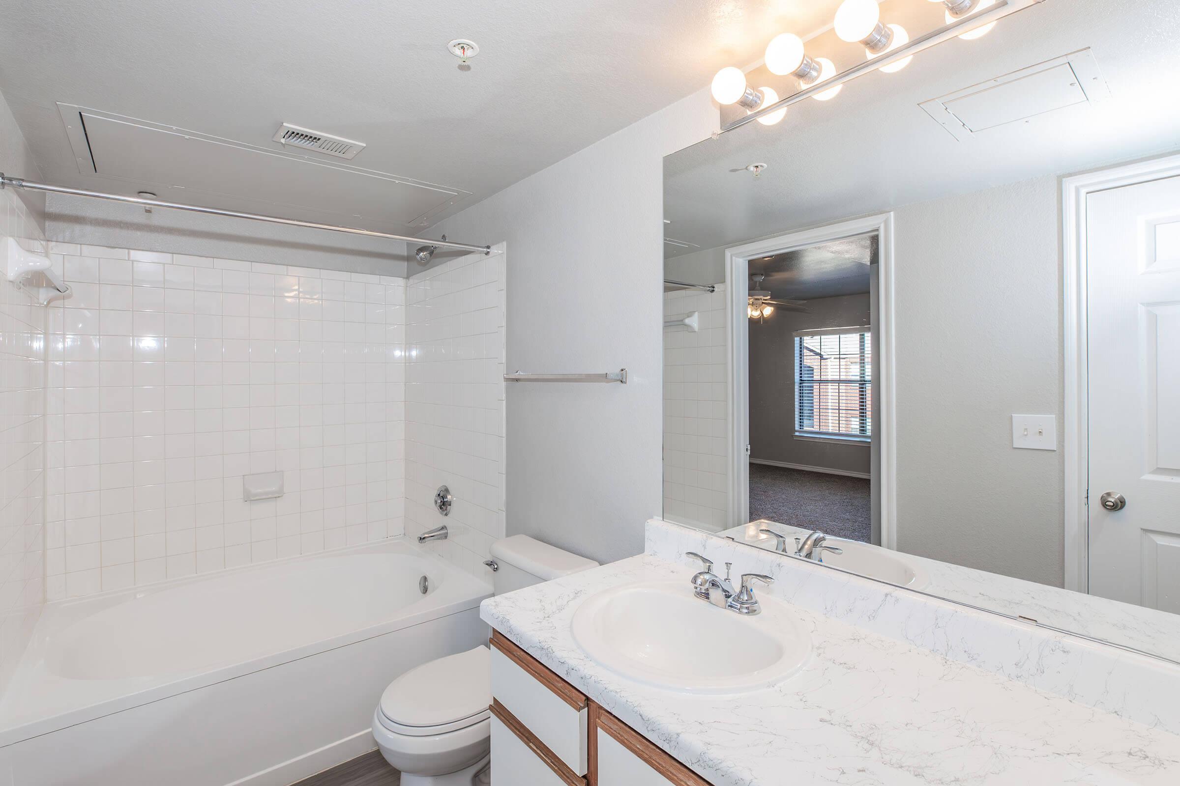 A bright, modern bathroom featuring a bathtub with a shower, a double-sink vanity with white marble countertops, and a large mirror. The walls are painted light gray, and a towel rack is visible. In the background, a glimpse of another room with a window and carpeted flooring can be seen.
