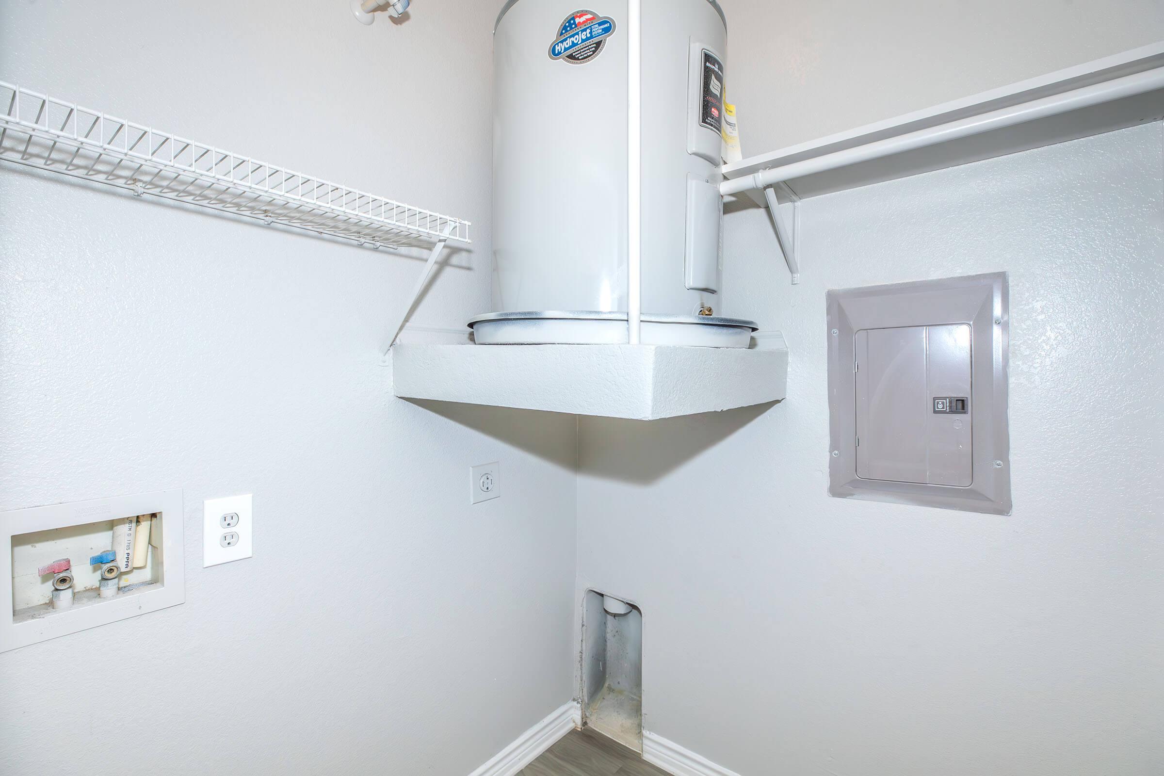A small utility room featuring a white water heater mounted on a concrete base, with a wire shelf above it. On one wall, there are electrical outlets and a circuit breaker panel. The walls are painted light gray, and the floor is tiled in a light color.