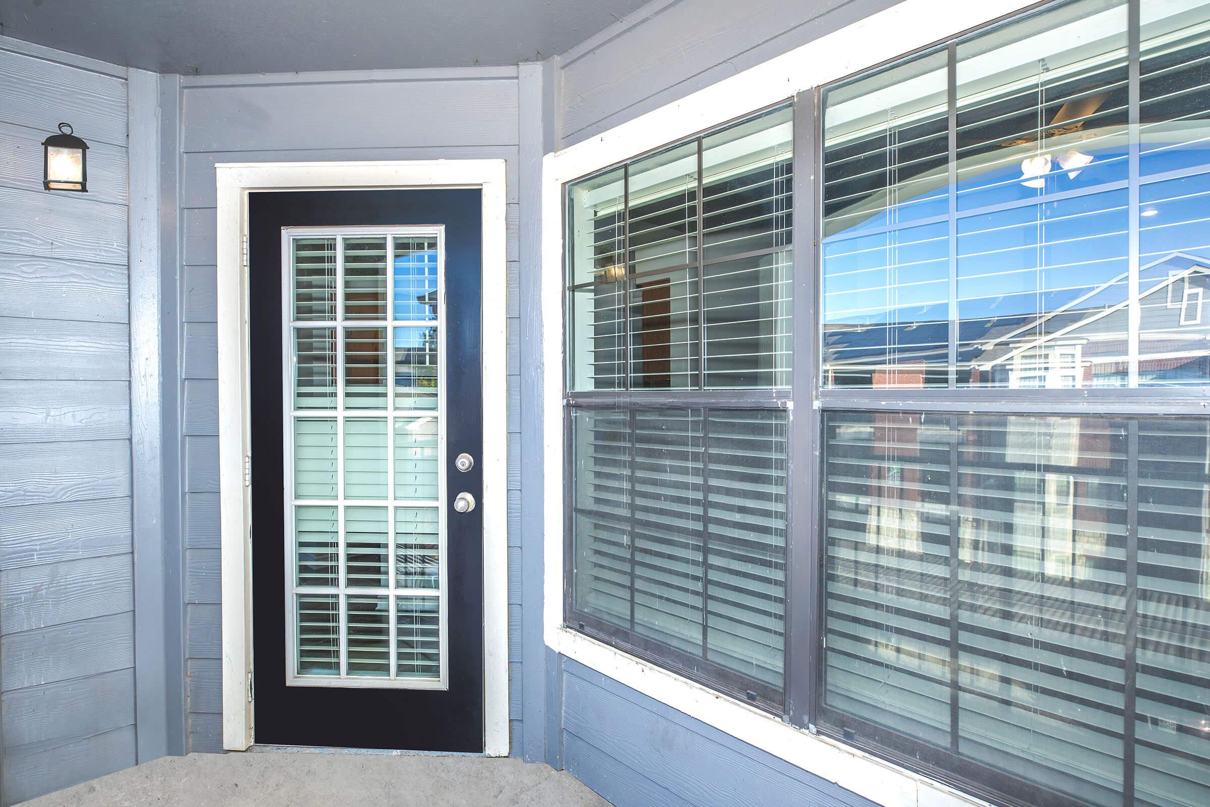 Exterior view of a porch featuring a black front door with a grid pattern of glass panes. Surrounding the door are light gray wooden walls, and there are large windows with horizontal blinds. The porch is partially covered, providing a welcoming entrance.