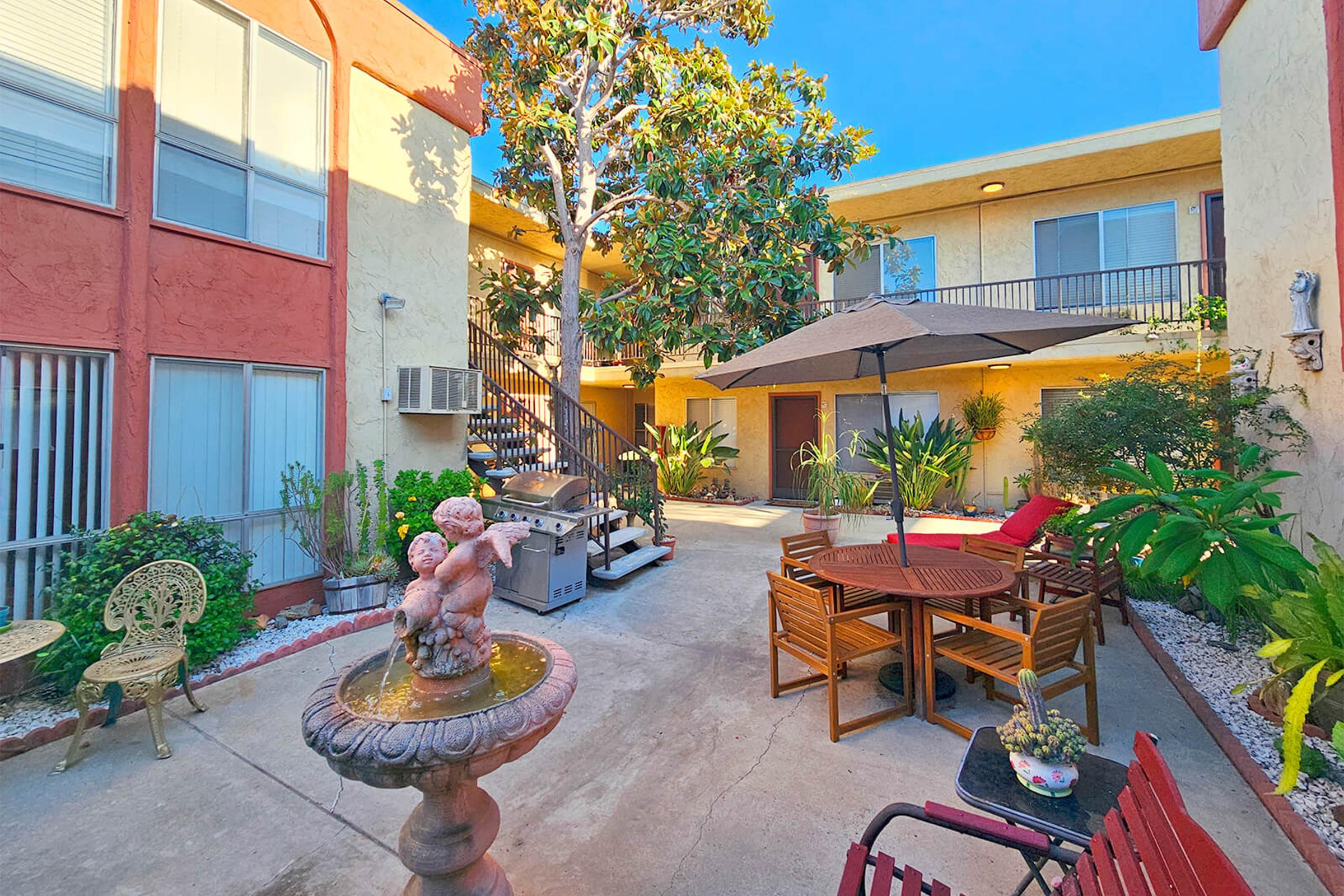 A sunny courtyard with a small fountain and a sculpture, surrounded by tropical plants and outdoor furniture. There are tables and chairs under an umbrella, along with a grill, set among the buildings with staircases leading to upper levels. The atmosphere is inviting and relaxed.