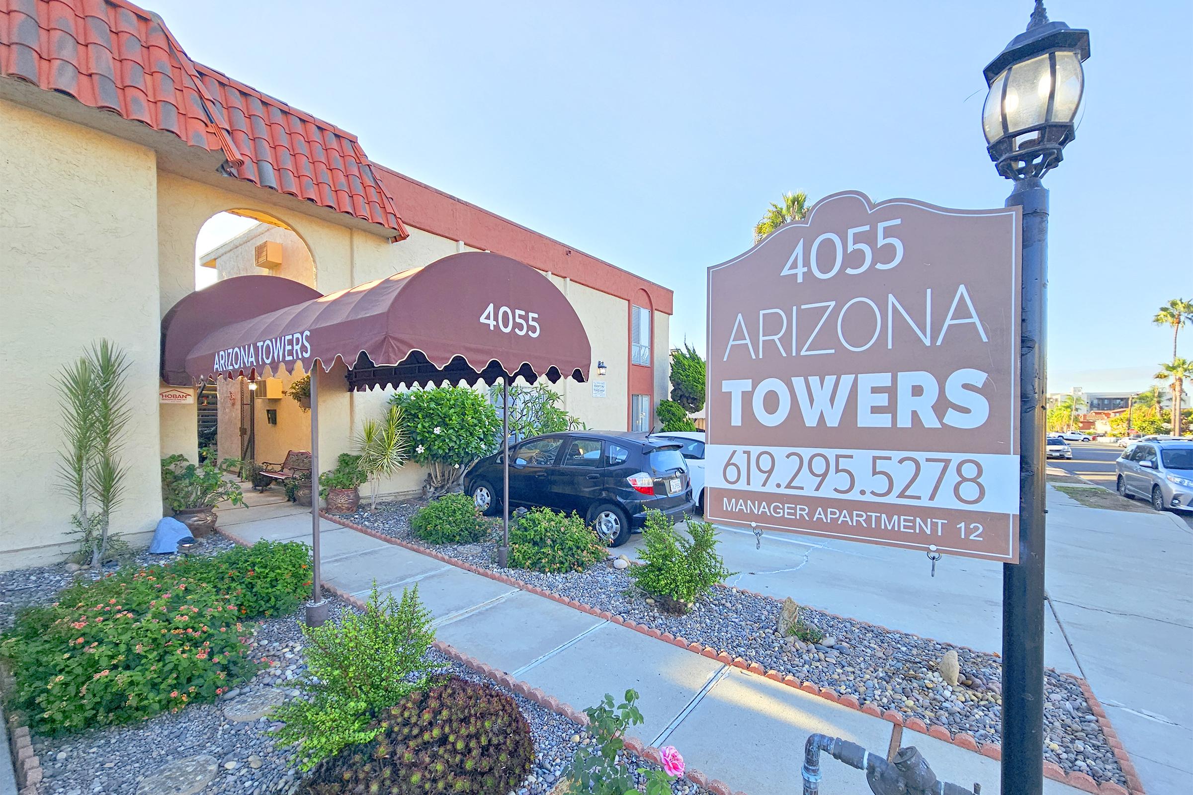 Sign for Arizona Towers located at 4055, with contact number 619-295-5278. The entrance features a covered awning and landscaped pathway. A parked car is visible in the driveway, with palm trees and shrubbery enhancing the outdoor appearance of the building.