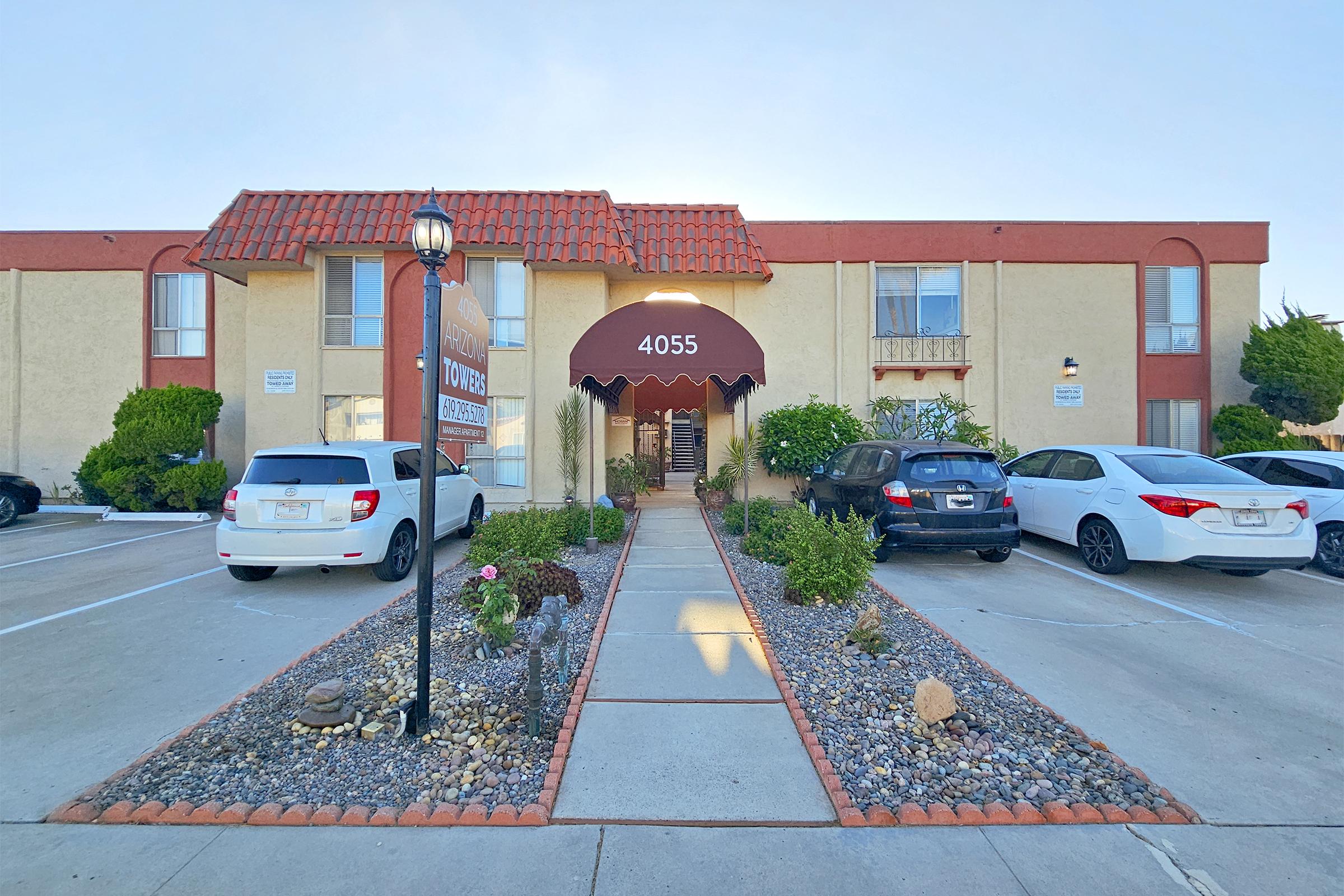 A front view of an apartment building featuring a numbered entrance "4055" under a maroon awning. The pathway leading to the entrance is lined with decorative rocks and plants. There are several parked cars on either side of the walkway, and the building has a light beige exterior with red accents.