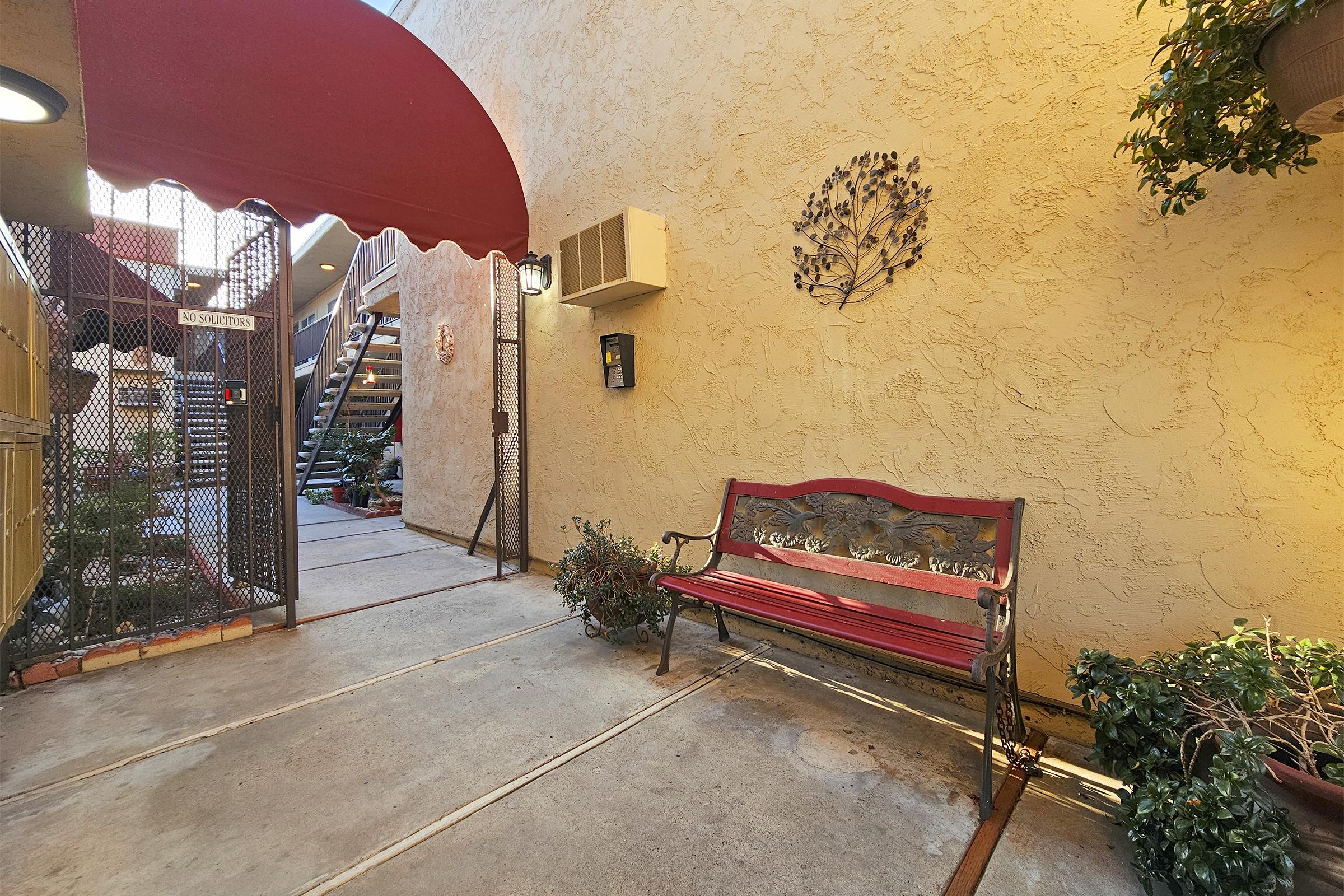 A cozy courtyard featuring a red bench against a textured beige wall. The entrance has a maroon awning, and there are potted plants nearby. A staircase is visible in the background, leading to upper levels, with a decorative wall hanging next to the entrance. Soft lighting creates a warm atmosphere.