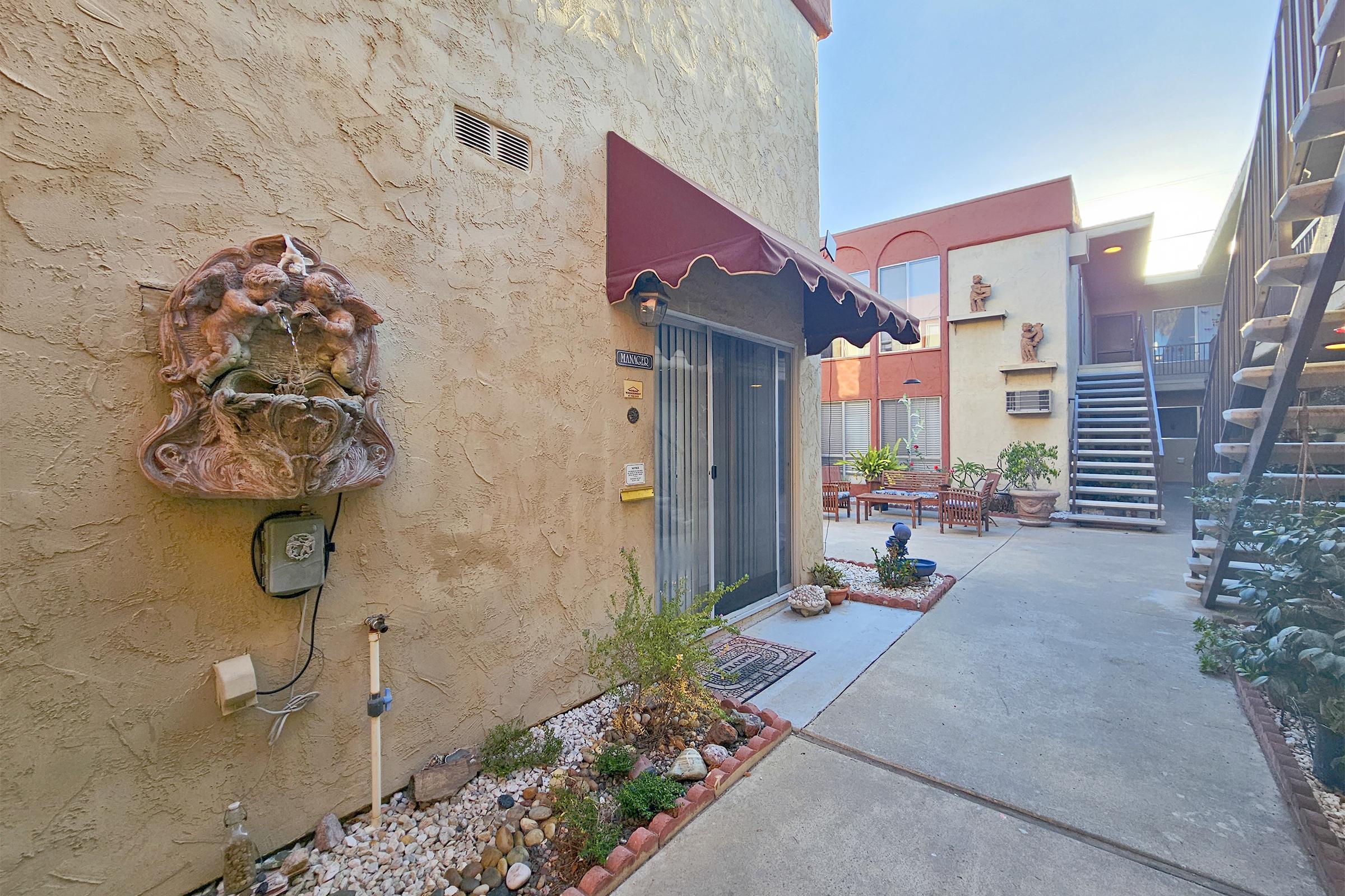 A courtyard view of a residential complex, featuring a decorative wall plaque, potted plants, and a small patio area with outdoor furniture. The scene is illuminated by soft sunlight, highlighting the warm tones of the building's exterior and the well-kept landscaping along the walkway.