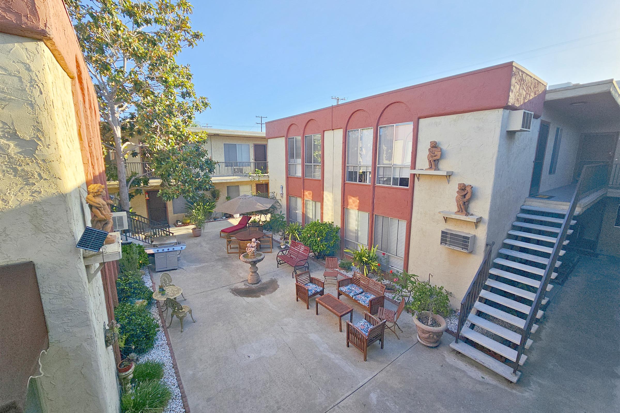 View of a courtyard in an apartment complex featuring outdoor furniture, potted plants, a decorative fountain, and sculptures. The buildings have red and beige exteriors, and there are stairs leading to upper levels. Sunlit space creates a welcoming atmosphere.