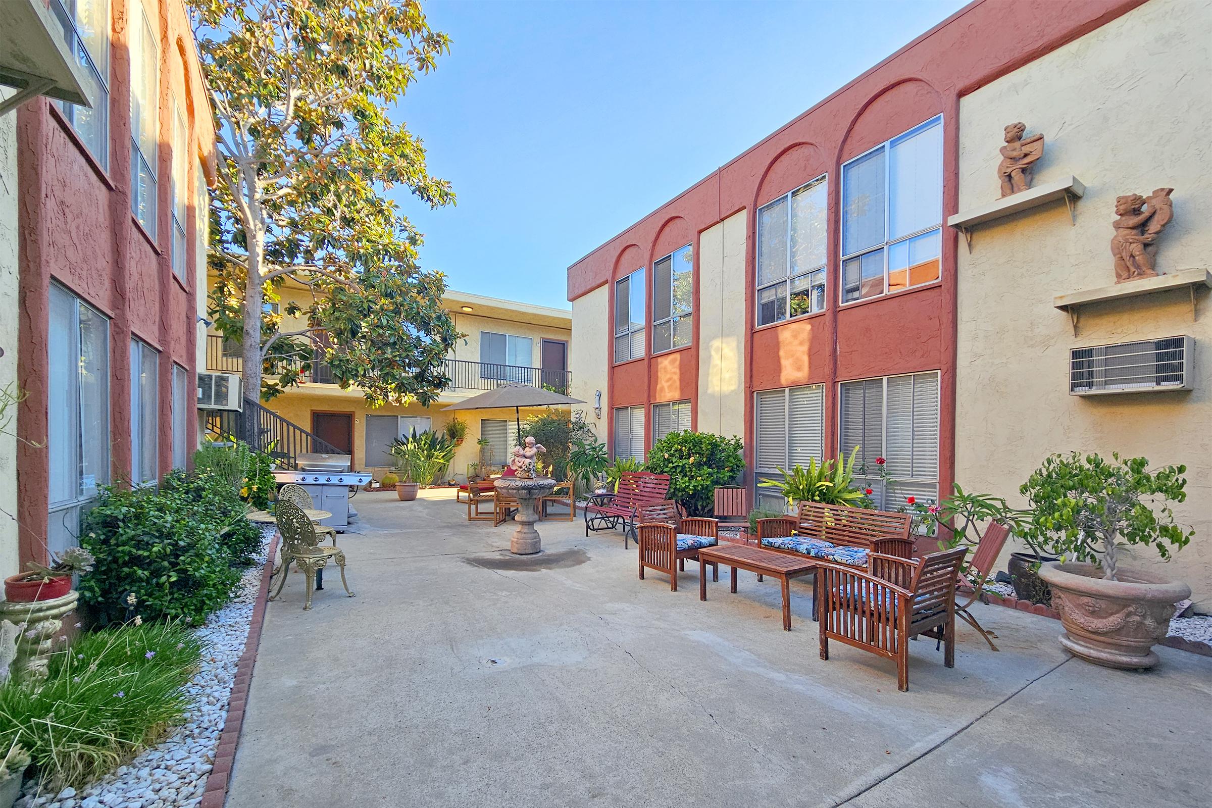 A sunny courtyard featuring terracotta-painted buildings with large windows, surrounded by greenery. Several wooden benches and chairs are arranged along a pathway, with potted plants adding to the outdoor ambiance. A bright blue sky creates a cheerful atmosphere in this welcoming space.