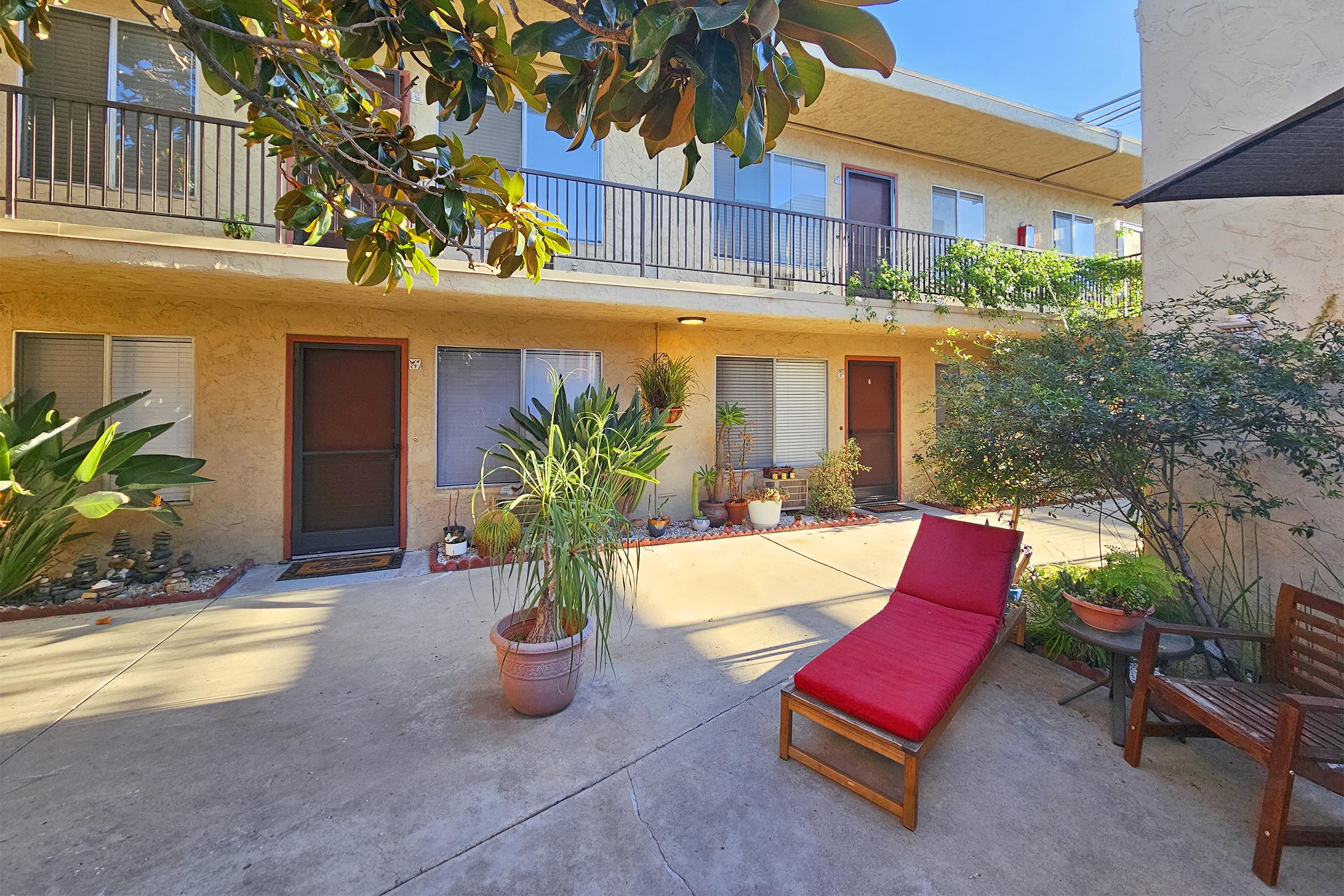 A view of a sunny courtyard in an apartment complex. It features a red lounge chair, potted plants, and a decorative pathway. The two-story building has balconies and several green plants, creating a relaxing outdoor space.