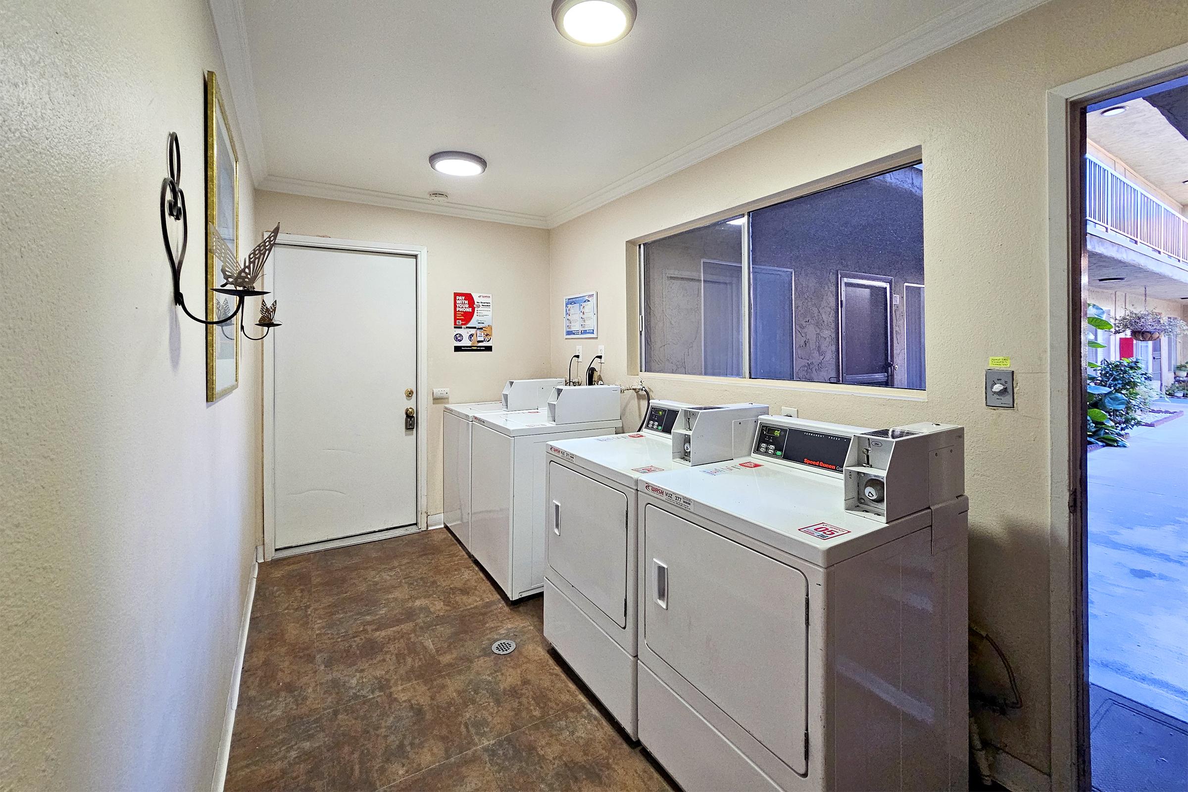 A laundry room featuring several white washing machines and dryers lined up against the wall. There's a door on one side, a window allowing natural light, and a simple light fixture on the ceiling. The floor is tiled, and the walls are painted a neutral color.
