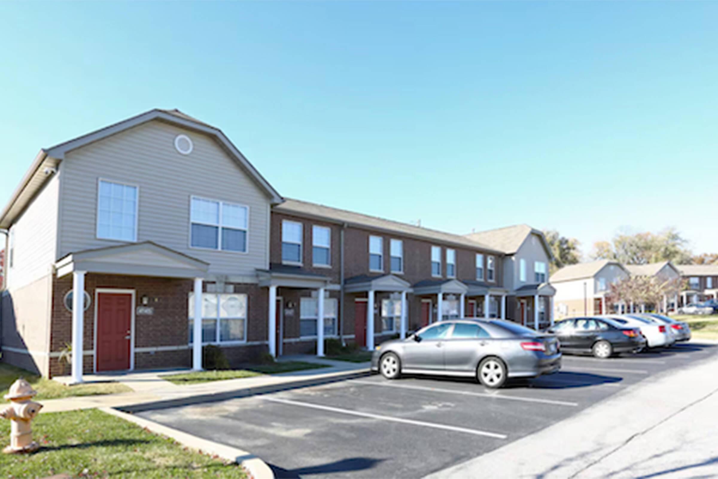 A row of two-story townhouses featuring beige and brown siding, with multiple parked cars in the foreground. The scene is set on a clear day with blue skies. There is a fire hydrant nearby, and the area is landscaped with grass.