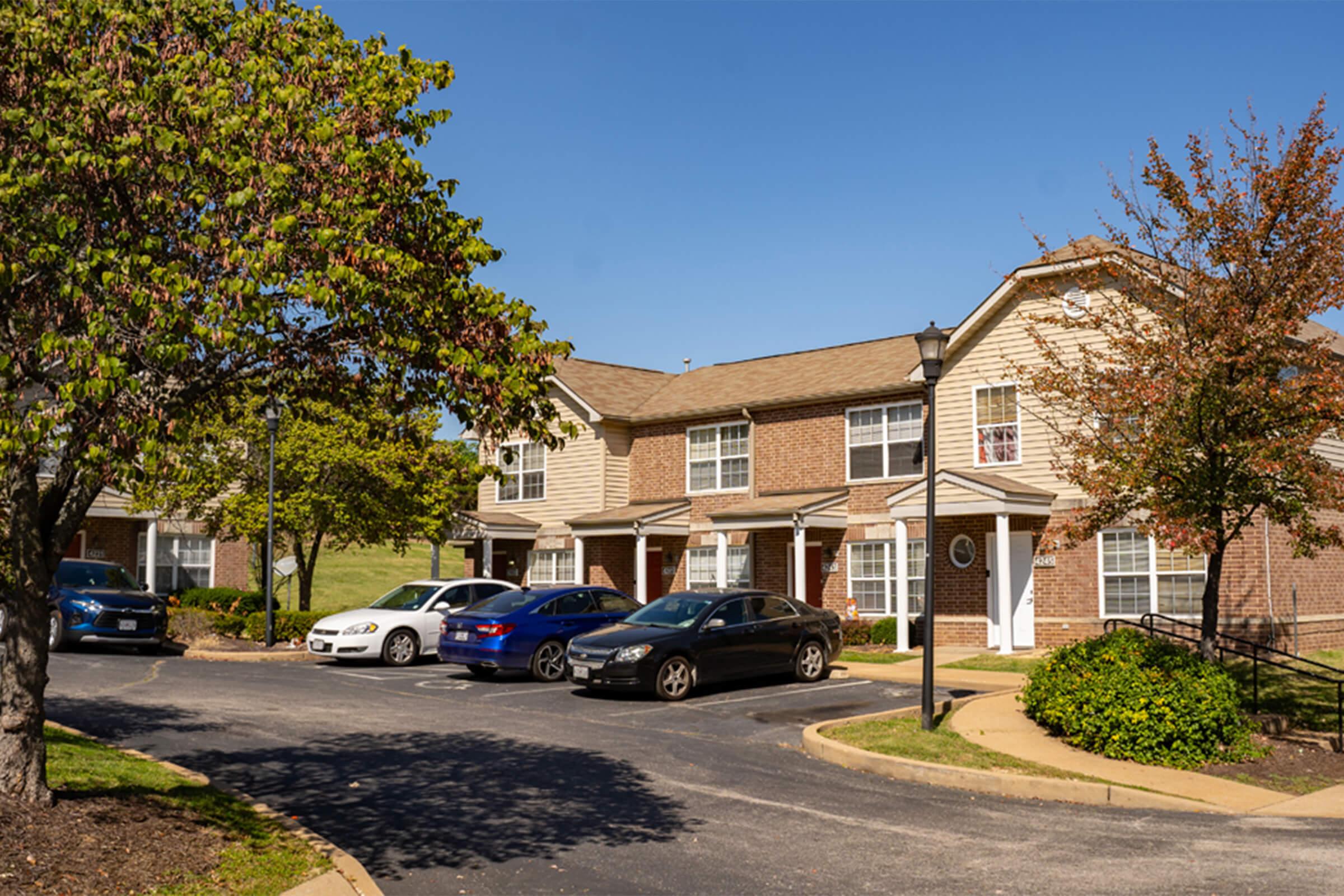 A row of two-story brick apartment buildings with well-maintained landscaping. Cars are parked in front of the units, and clear blue skies are visible in the background. Green trees and shrubs enhance the outdoor setting, creating a pleasant residential atmosphere.