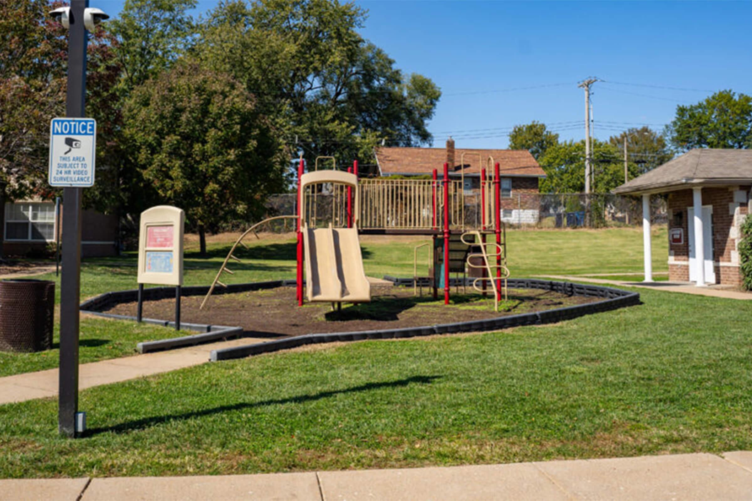 A playground featuring a colorful play structure with slides, climbing features, and swings, surrounded by grassy areas and a paved pathway. There is a notice sign nearby, and trees provide shade in the background, creating a welcoming outdoor space for children.