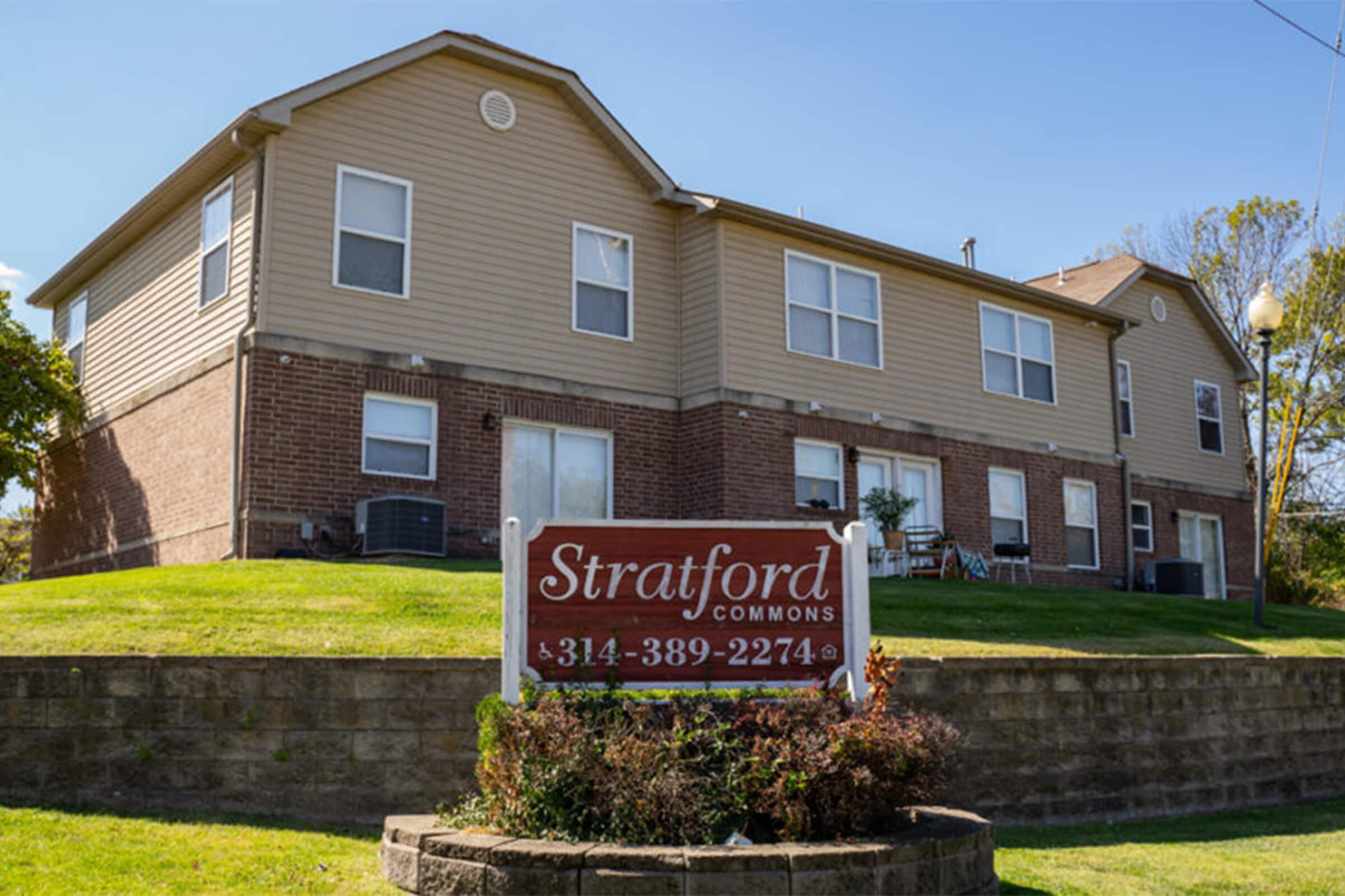 A two-story apartment building with brick and beige siding. The building has several windows and a sign in front that reads "Stratford Commons" along with a contact number. The property features a well-maintained lawn and landscaping. Clear blue sky in the background.