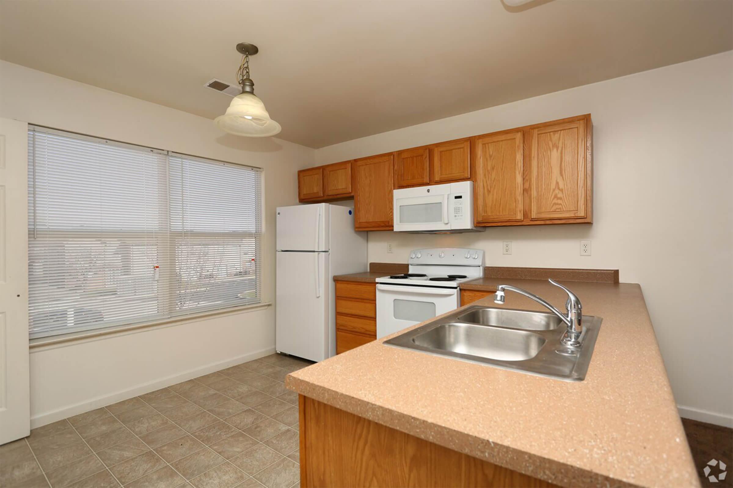 A modern kitchen featuring wooden cabinets, a white refrigerator, a microwave above the stove, and a sink. The counter has a light brown surface, and there's a large window allowing natural light to fill the room. The floor has light-colored tiles, enhancing the spacious feel of the kitchen area.