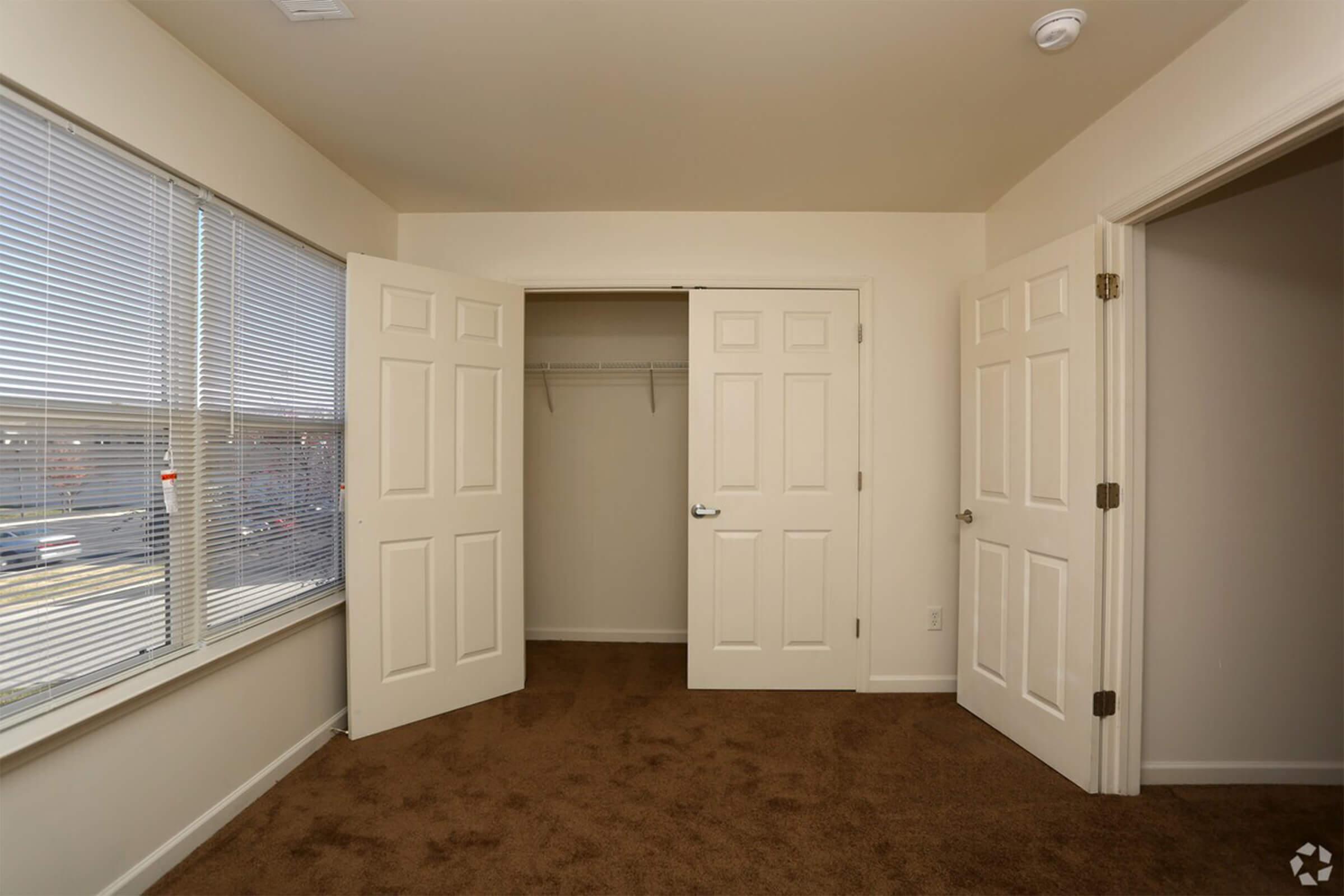 A small interior room with beige walls and brown carpet. Two doors are open to reveal a closet with a hanging rod. A large window on the left features horizontal blinds, allowing natural light to enter. The room appears empty and well-lit, offering a clean, minimalistic space.