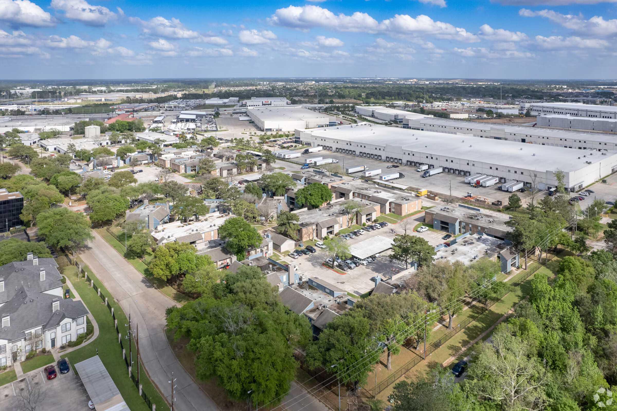 Aerial view of a suburban area featuring a mix of industrial buildings and residential neighborhoods. The scene includes tree-lined streets, warehouses in the background, and patches of greenery, under a partly cloudy sky. The layout shows a blend of urban development and natural elements.