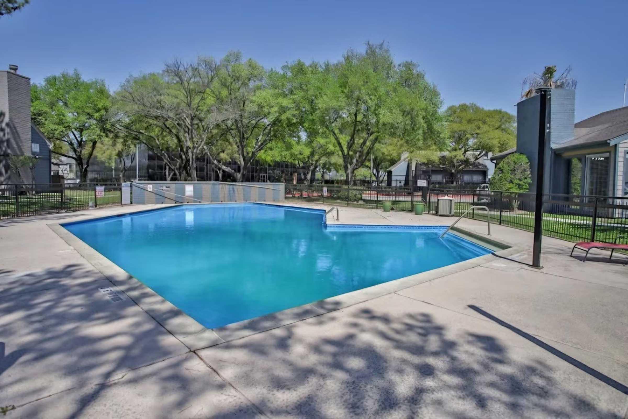 A clear blue swimming pool surrounded by a fence, with trees in the background and lounge chairs on the patio. The pool area is well-maintained, providing a serene environment for relaxation. The sky is clear and sunny, enhancing the inviting atmosphere.