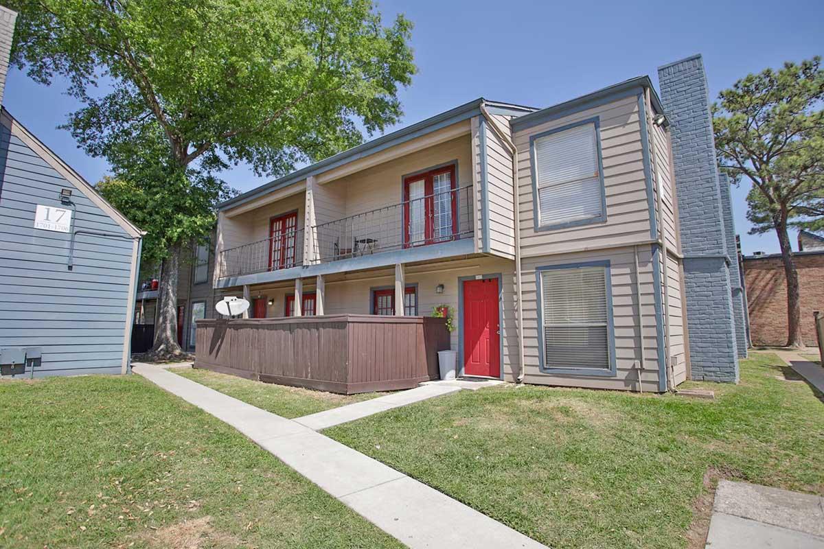 Two-story apartment building with gray siding and red doors. The balcony features black railings. In front, there is a small lawn with green grass and a white pathway leading to the entrance. Trees are visible in the background, and an adjacent blue building is partially visible on the left.
