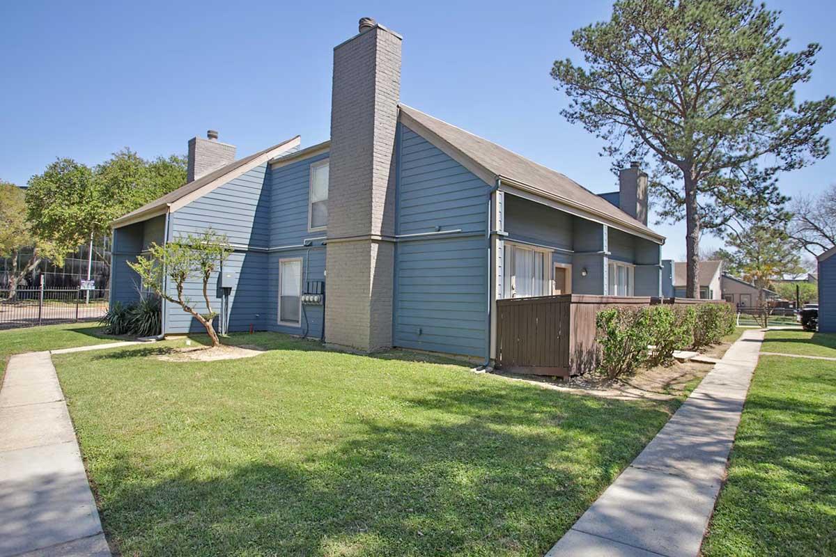 Two-story blue residential building with a sloped roof. The building features several windows, a chimney, and is surrounded by green grass and small trees. A sidewalk runs alongside the building, leading to a fenced area. Bright blue sky above completes the scene.